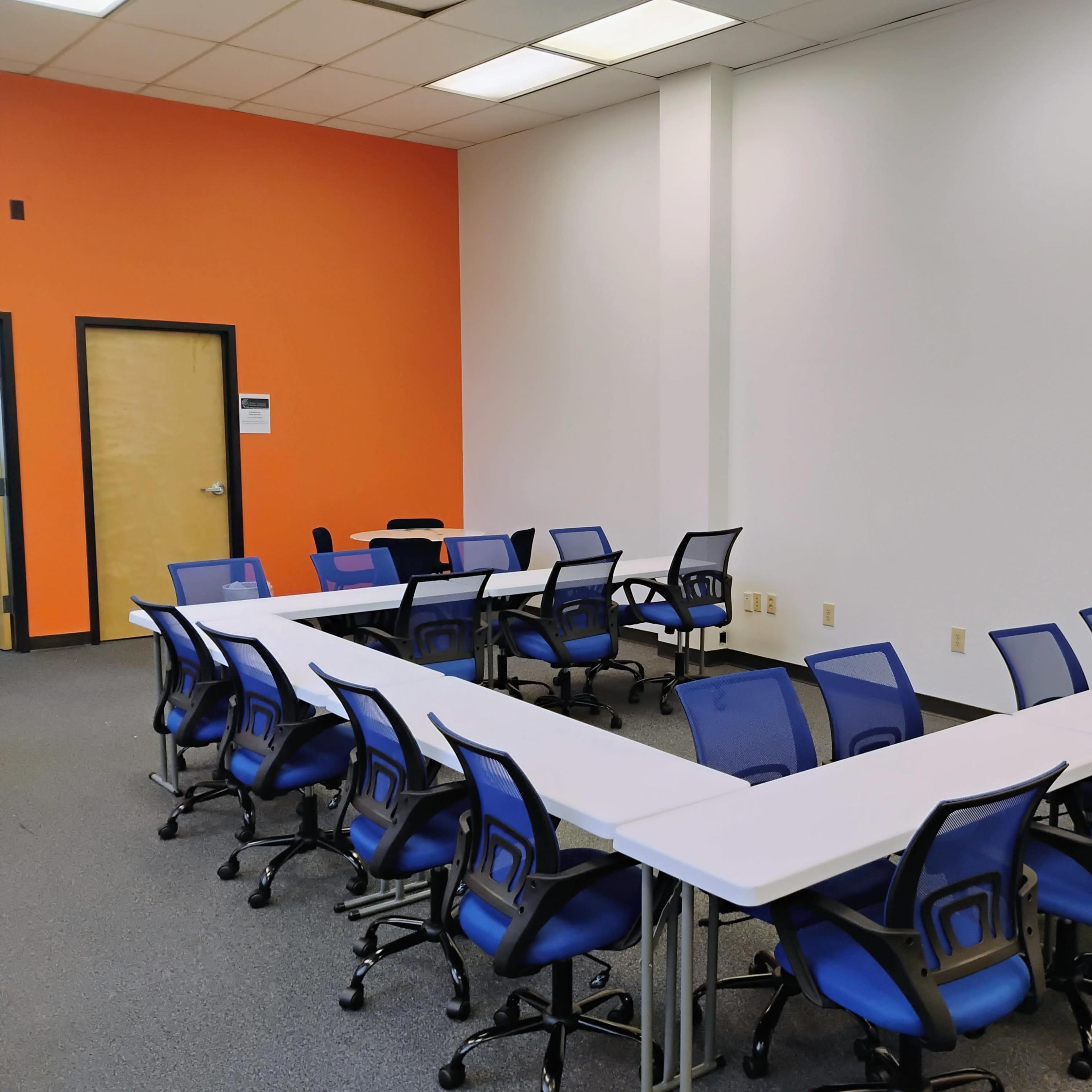 A conference room features multiple blue rolling chairs arranged around white tables in a U-shape against an orange wall.