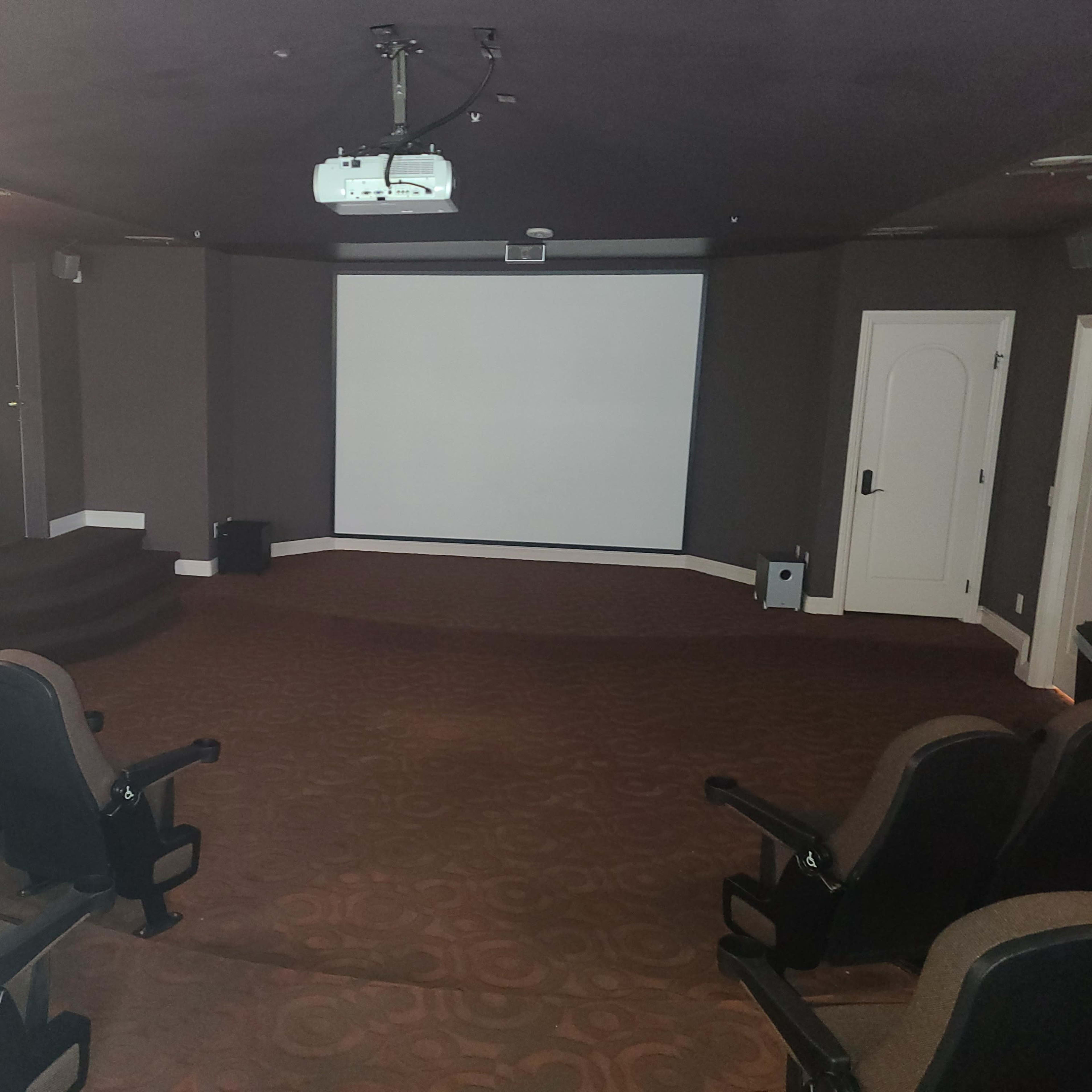 A private home theater featuring a blank screen at the front, tiered seating rows, and a projector mounted on the ceiling.