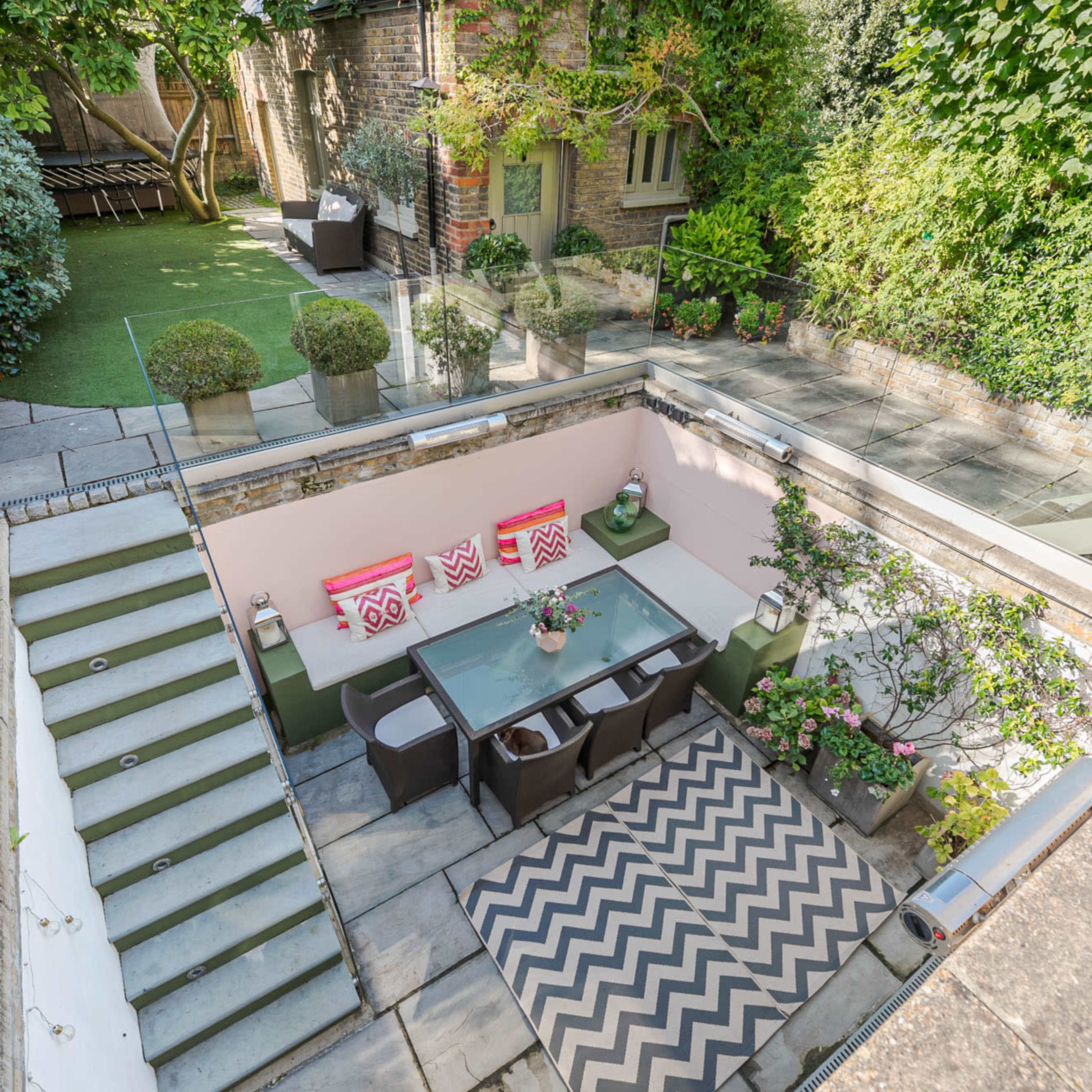 A sunlit outdoor dining area with a glass table surrounded by chairs, set on a patterned rug, and framed by lush greenery and a staircase leading to a garden.