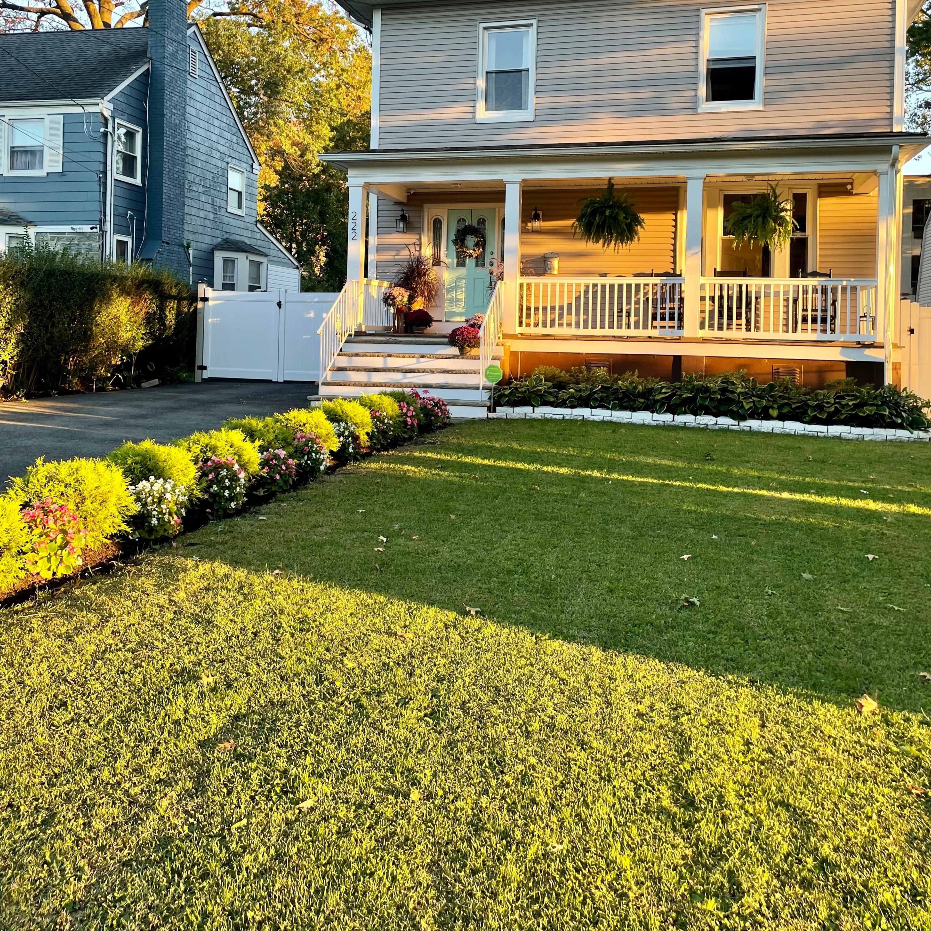 A three-story gray house with a front porch is surrounded by well-maintained lawns and flower beds.