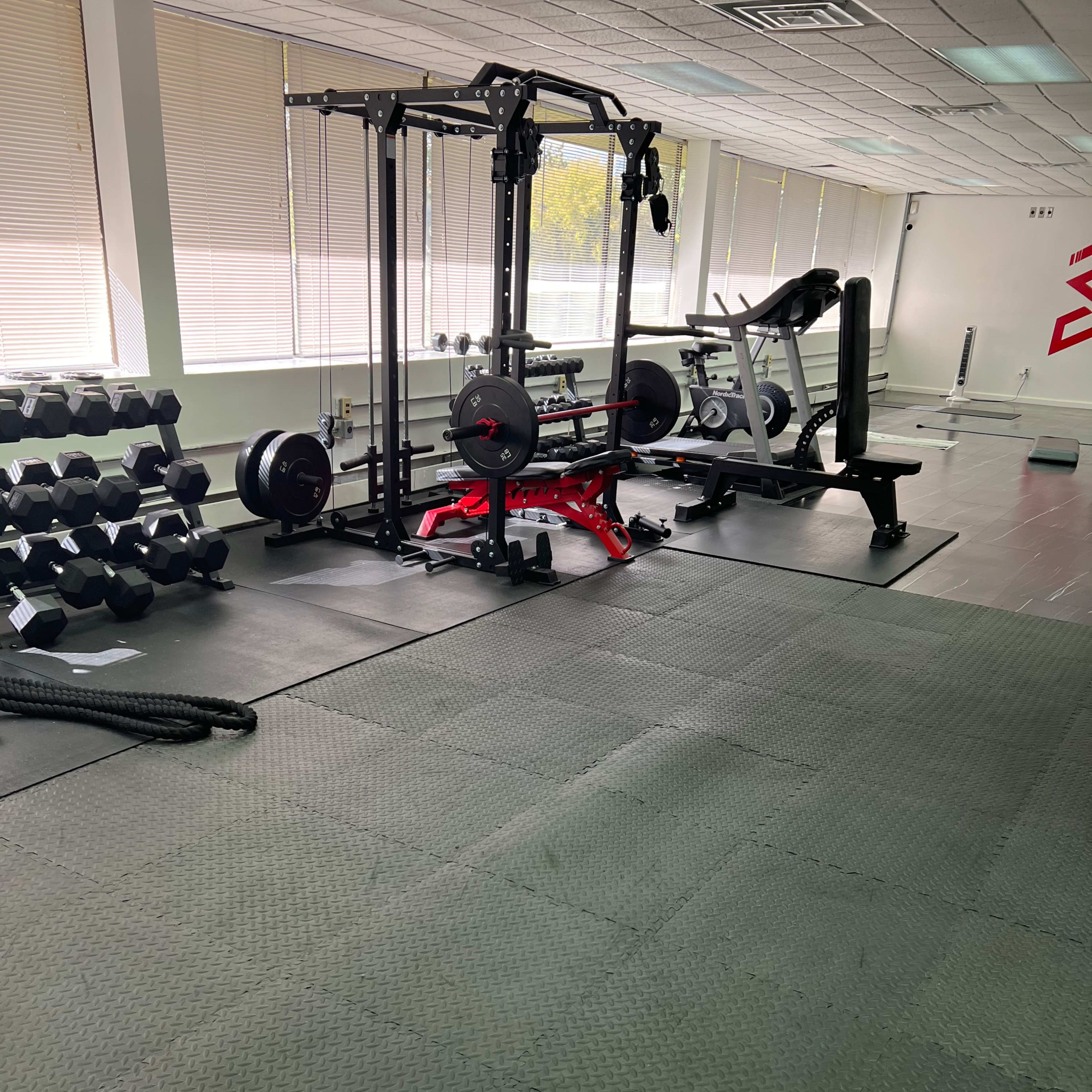 A gym interior with weightlifting equipment, including a squat rack and dumbbells, arranged on a rubber mat flooring.