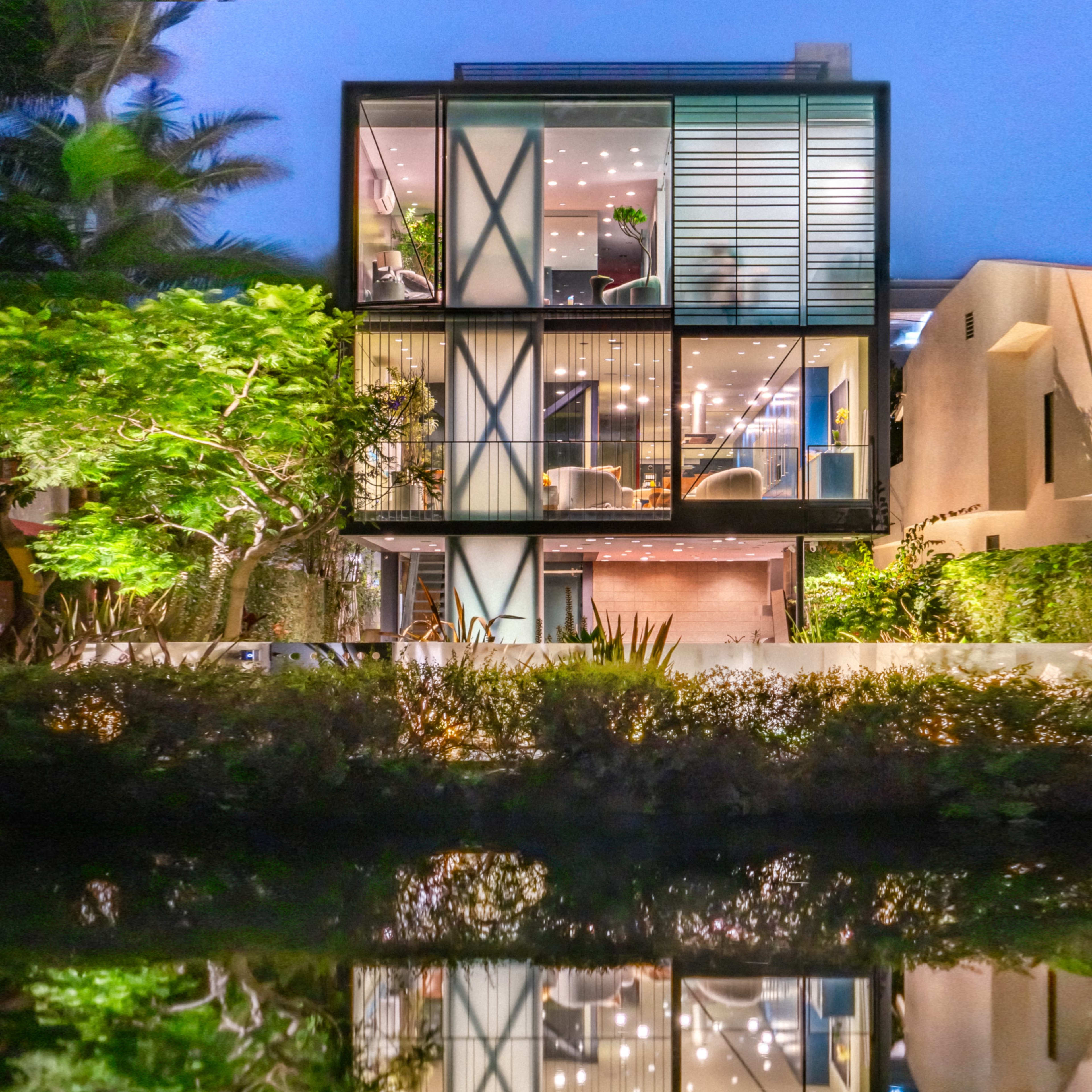 A modern three-story house featuring large windows and a symmetrical design reflects in a calm body of water surrounded by greenery.