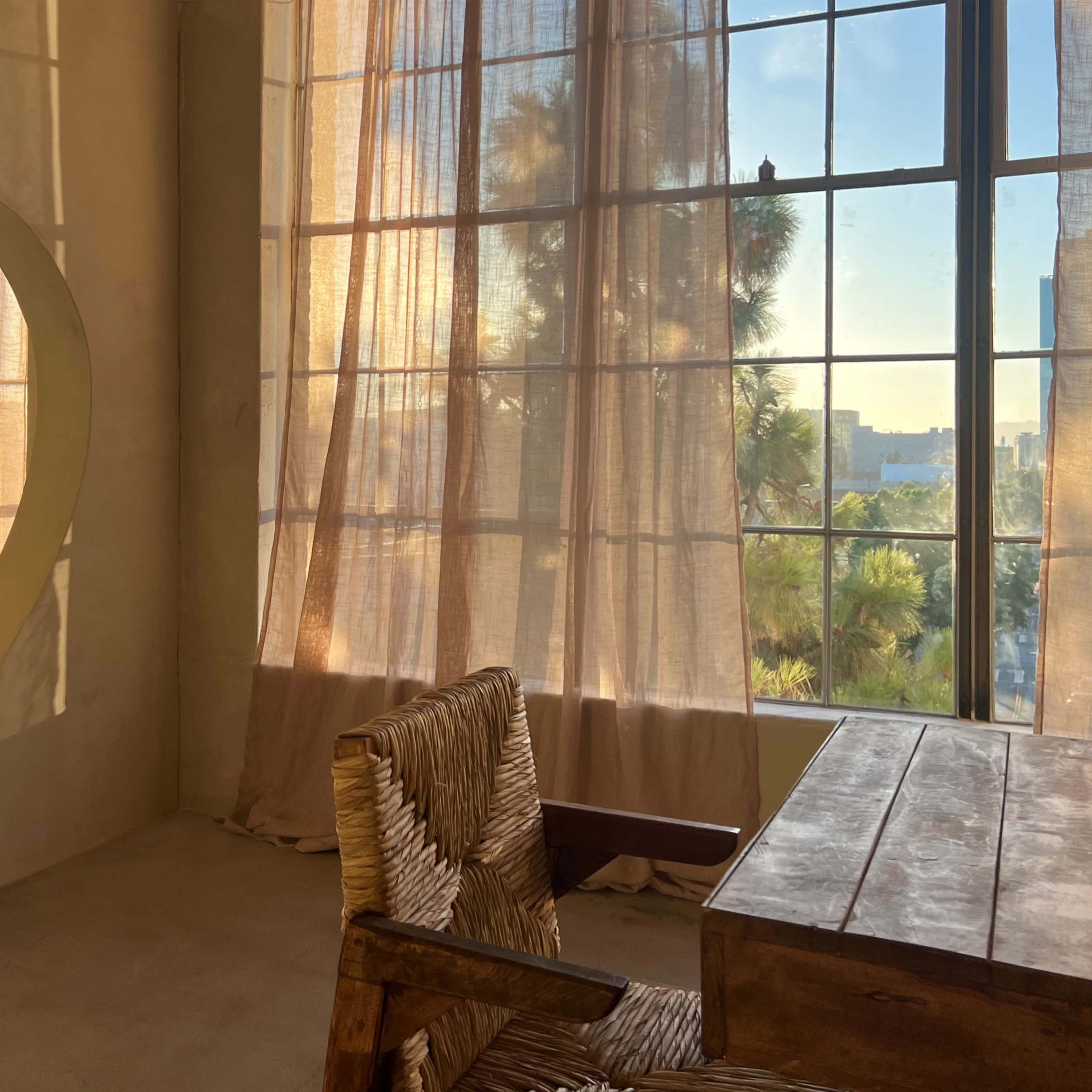 A room with large windows covered by sheer curtains and a round cutout reflecting light, featuring a wooden desk and woven chair.