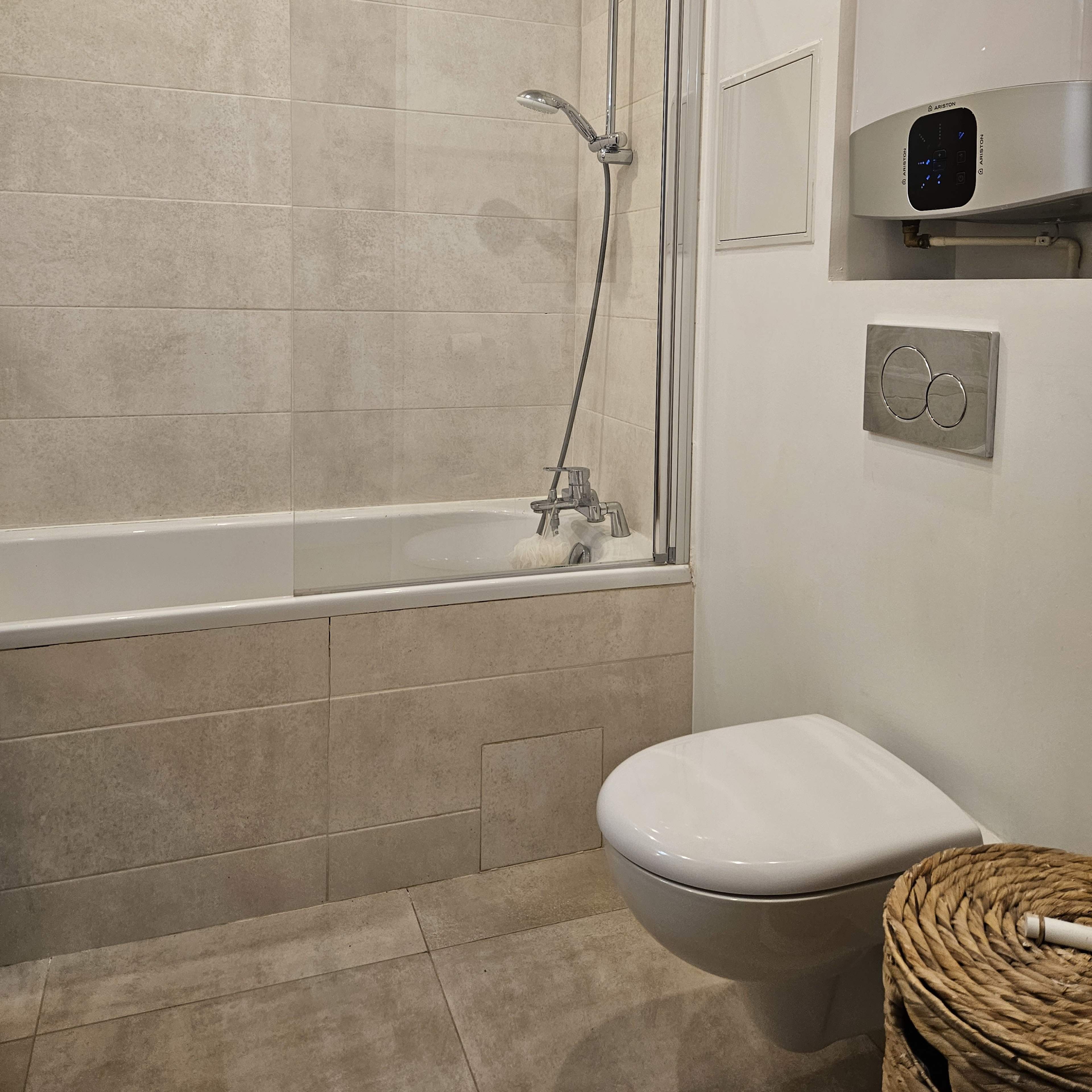 A modern bathroom featuring a bathtub with a shower, a wall-mounted toilet, and a light-colored tiled wall.