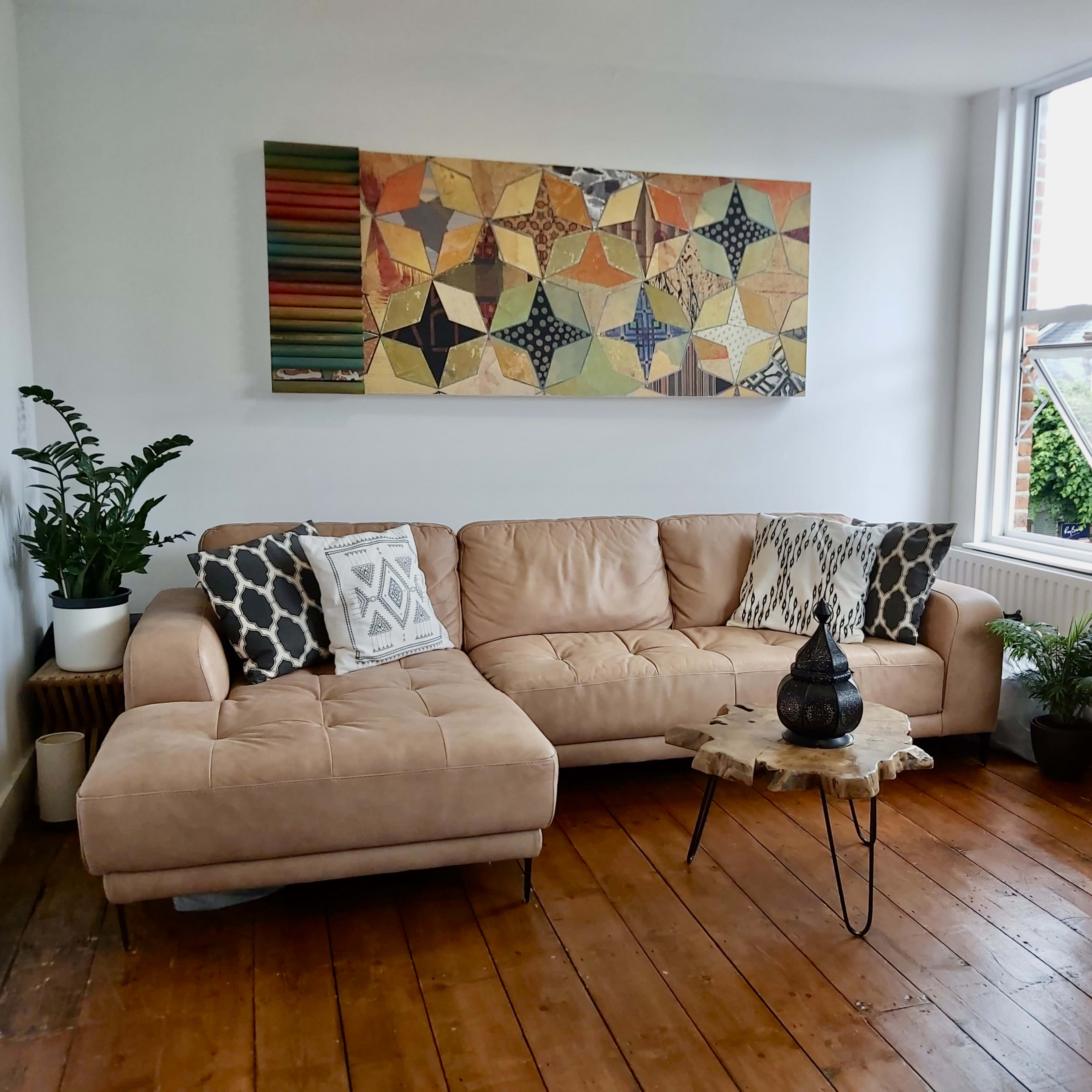 A cozy living room with a light-colored sectional sofa, a colorful wall art piece above it, and various plants in decorative pots.