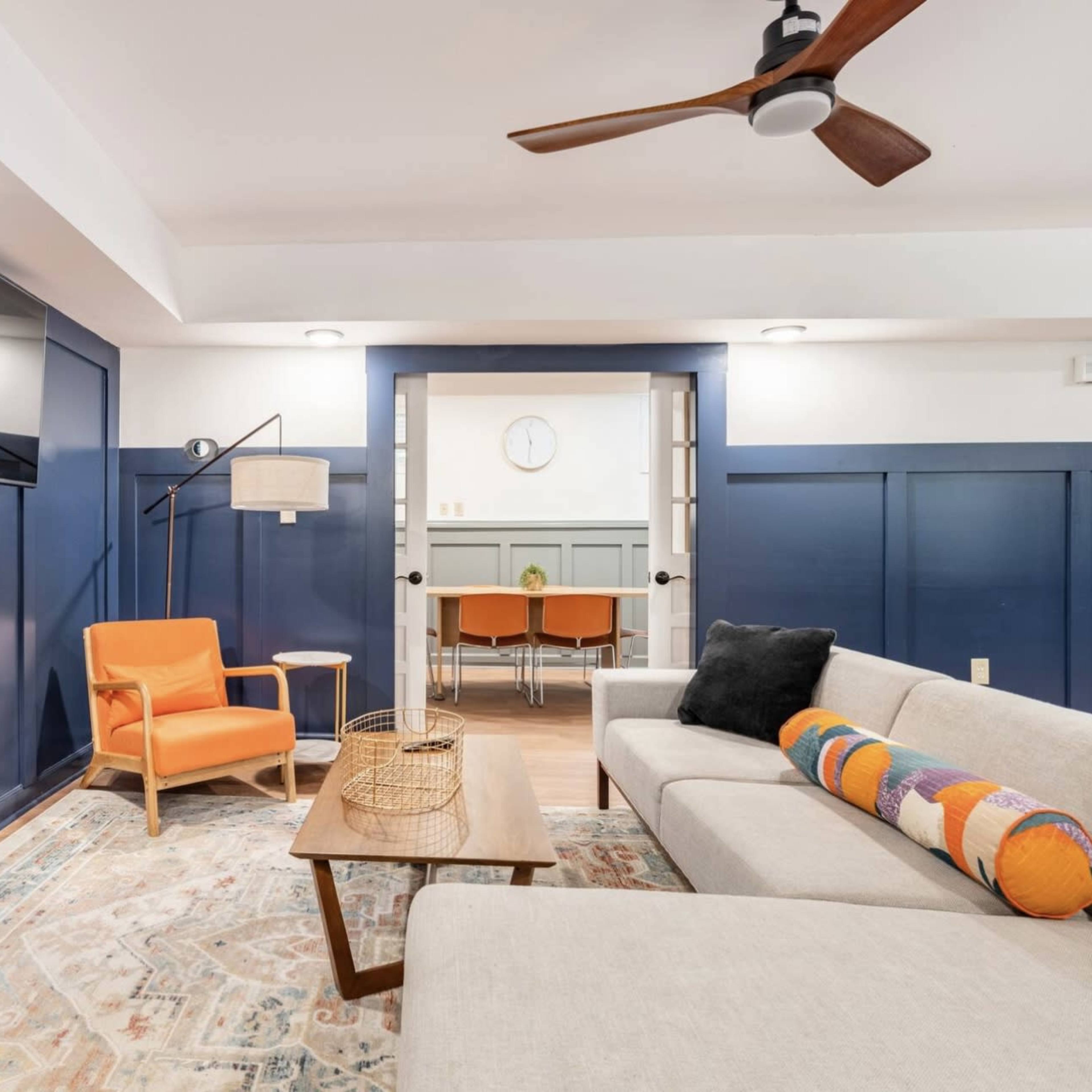 A living room with a sectional sofa, an orange armchair, a wooden coffee table, a flat-screen TV mounted on the wall, and a door leading to a dining area.