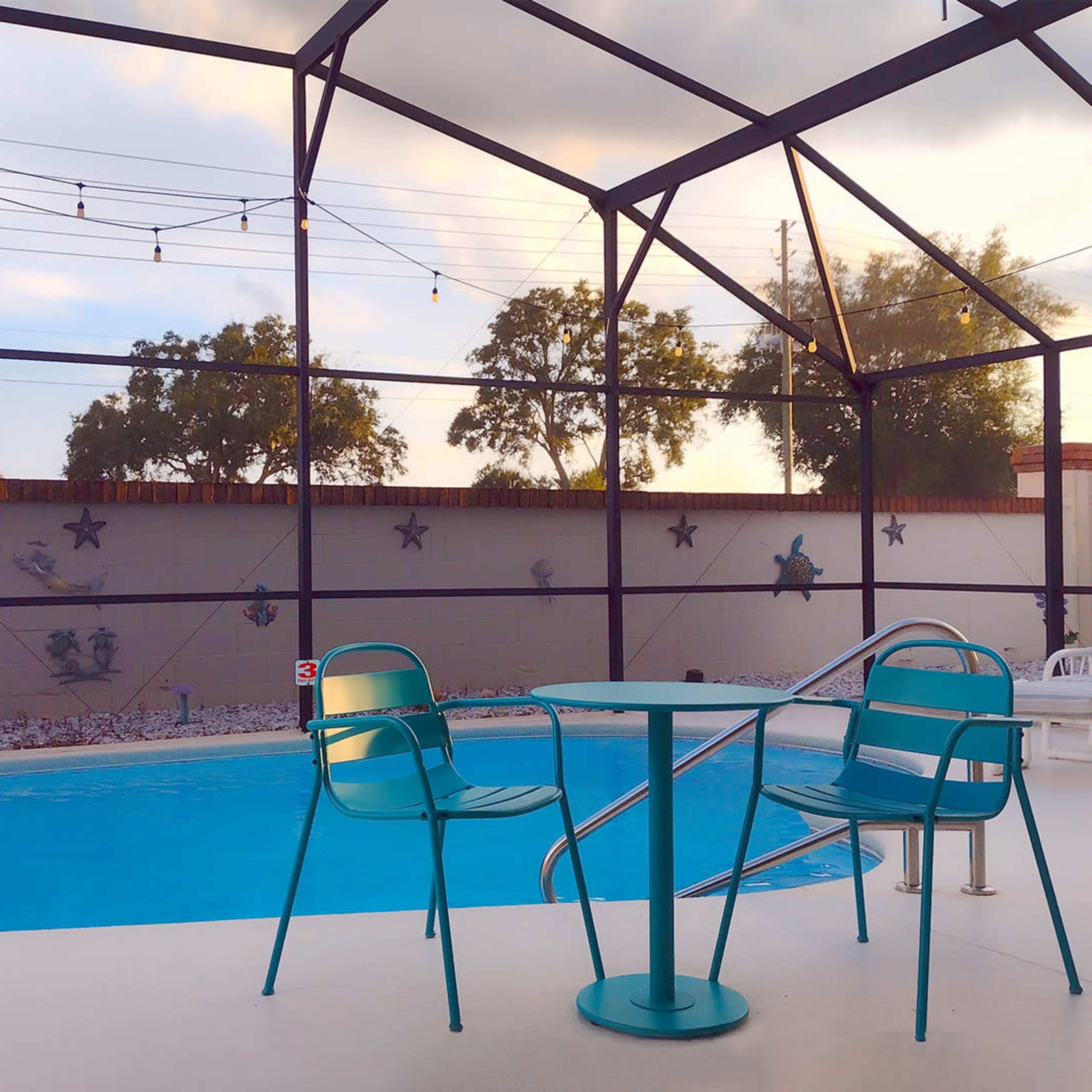A turquoise table and chairs sit beside a pool enclosed by a screen, with trees and a wall in the background.