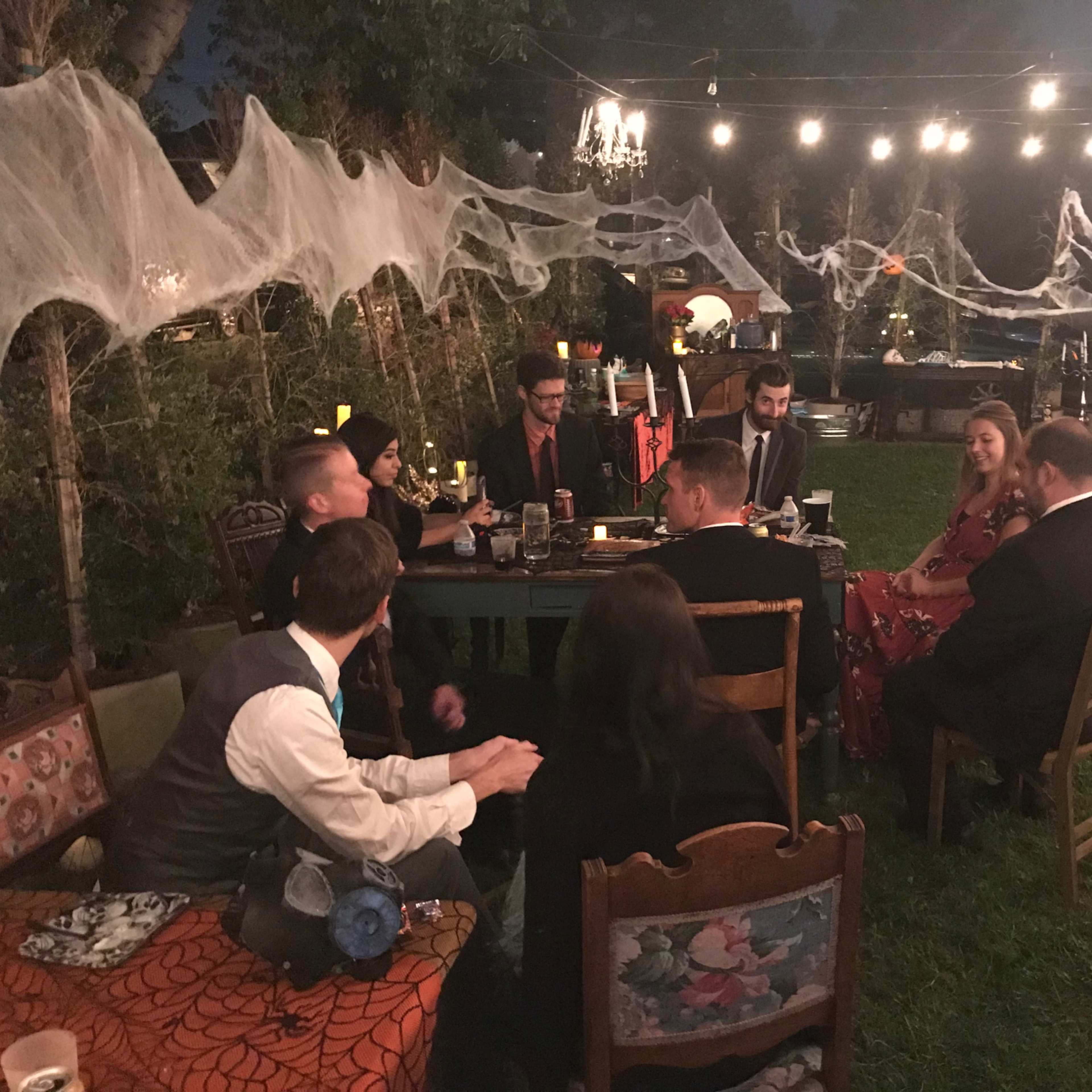 A group of people dressed in formal attire gather around tables decorated with Halloween-themed elements in a backyard setting.