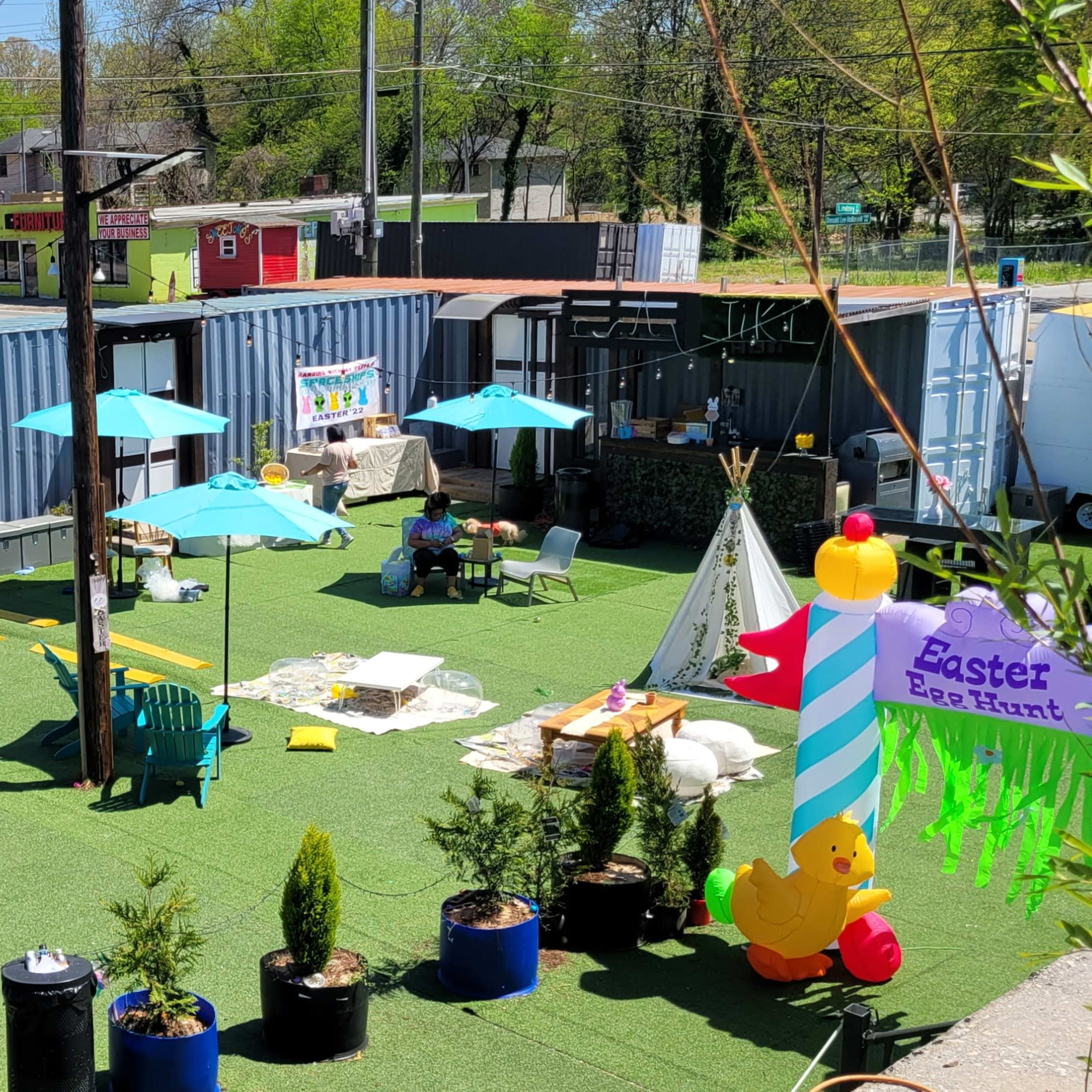 An outdoor event space with several tables arranged on green turf, colorful umbrellas, and decorations for an Easter egg hunt.
