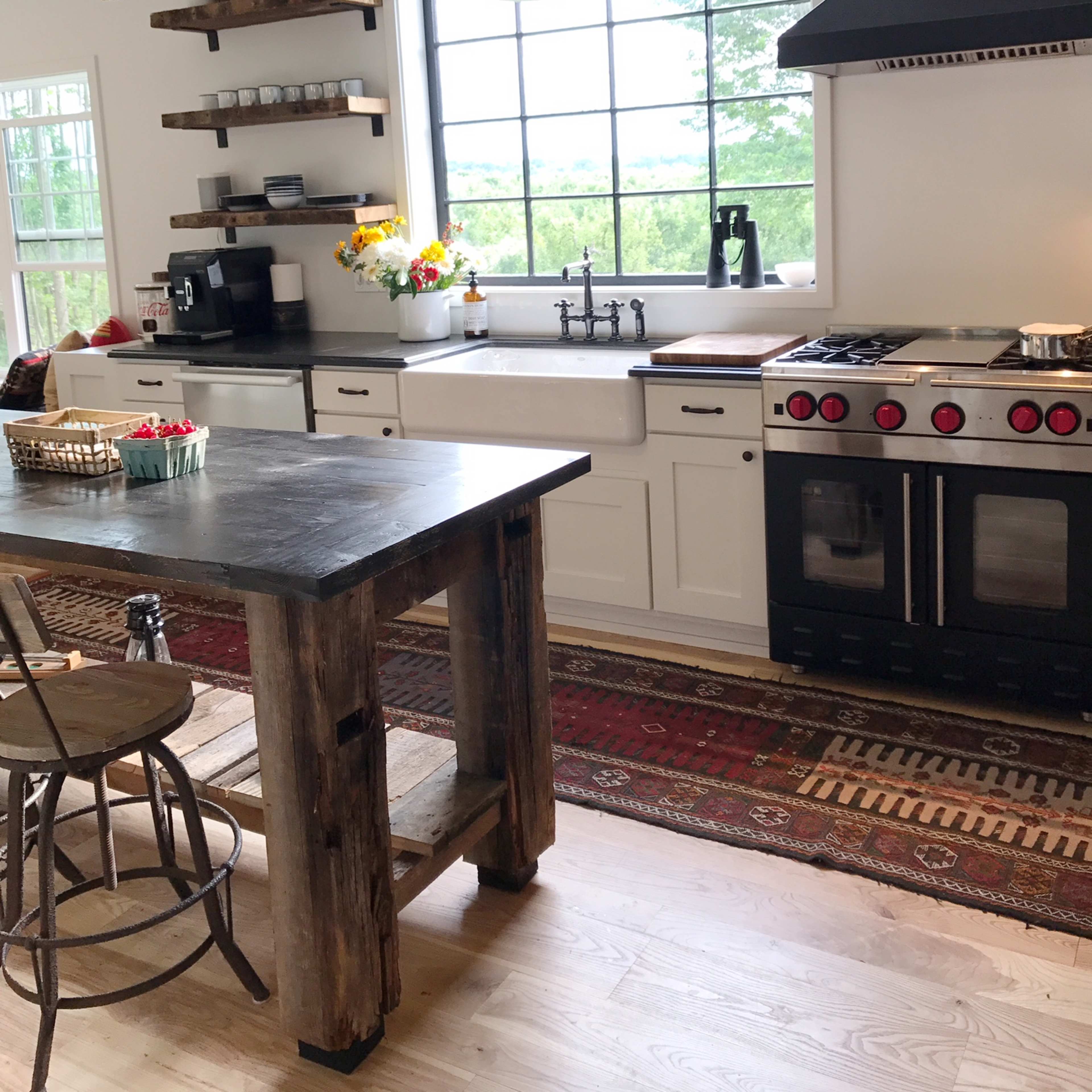 The kitchen features a central island made of wood, a large window overlooking greenery, and a modern stove with red knobs.