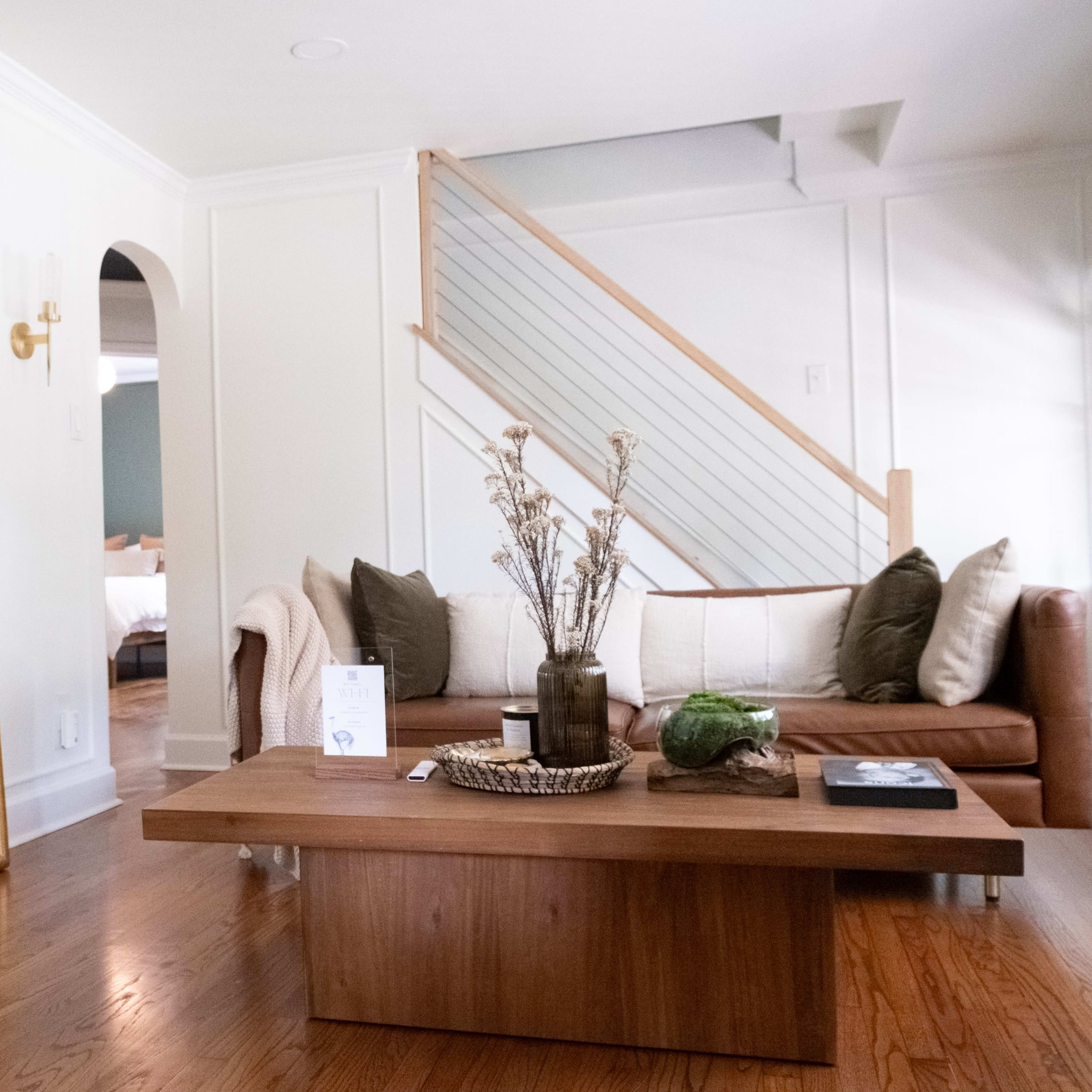 A well-decorated living room features a brown leather sofa, a wooden coffee table, and a large mirror reflecting a staircase.