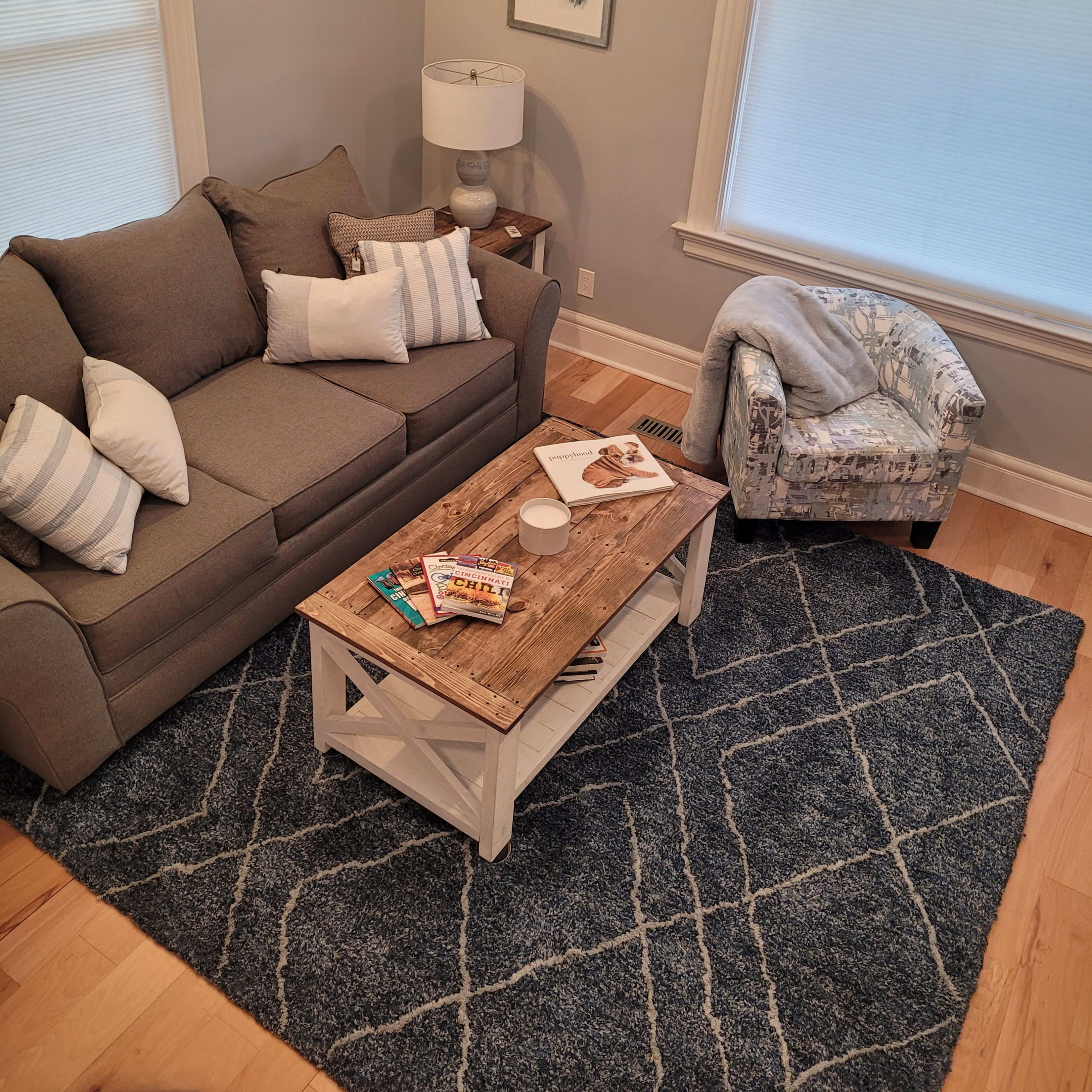A cozy living room with a grey couch, a wooden coffee table topped with magazines, and a patterned armchair on a blue area rug.