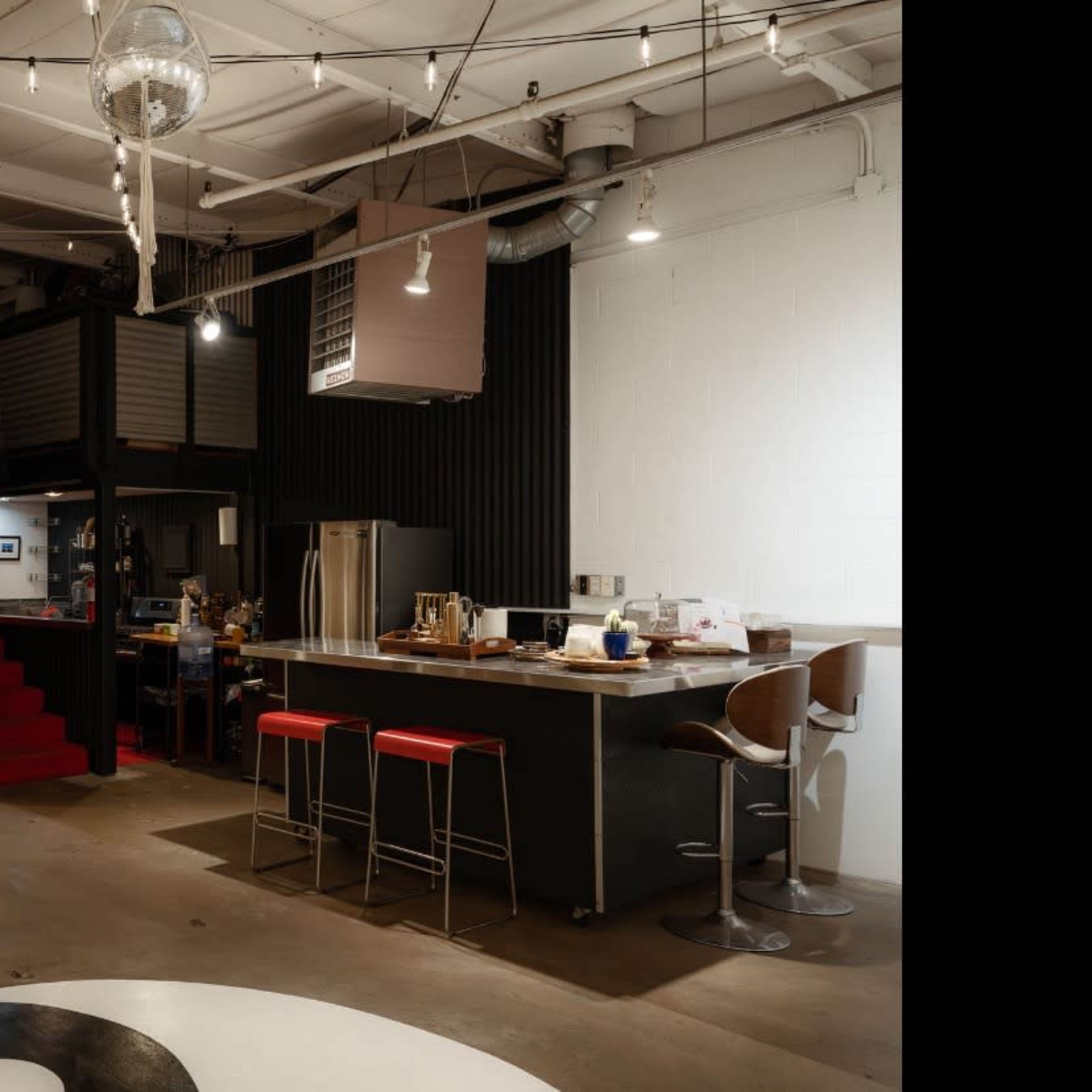 An industrial-style open-concept living space featuring a kitchen bar with red stools, a living area with a green couch, and a lofted section accessible by stairs.