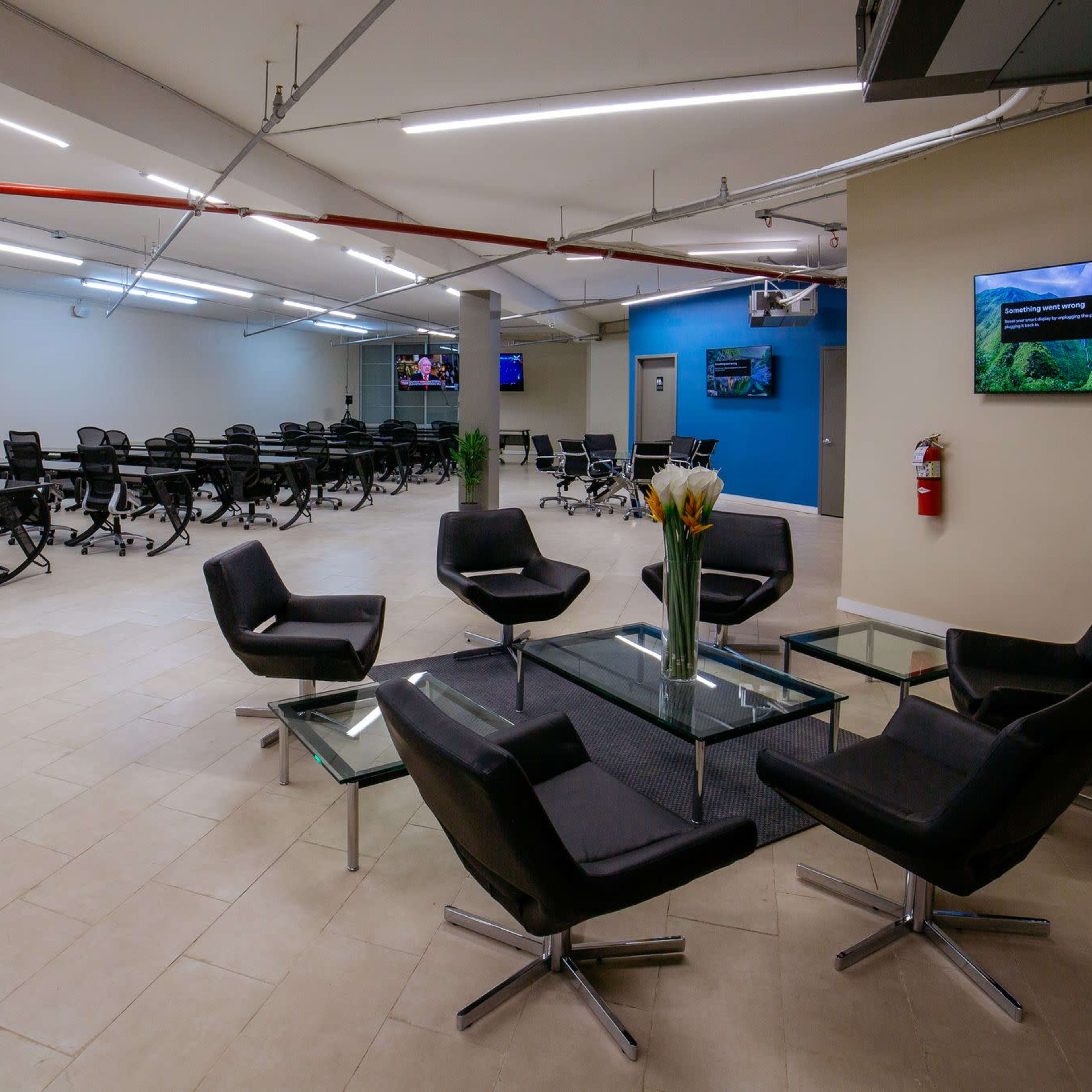 A modern office space with a seating area featuring black chairs around a glass table, and a large room in the background filled with empty office chairs and screens on the walls.
