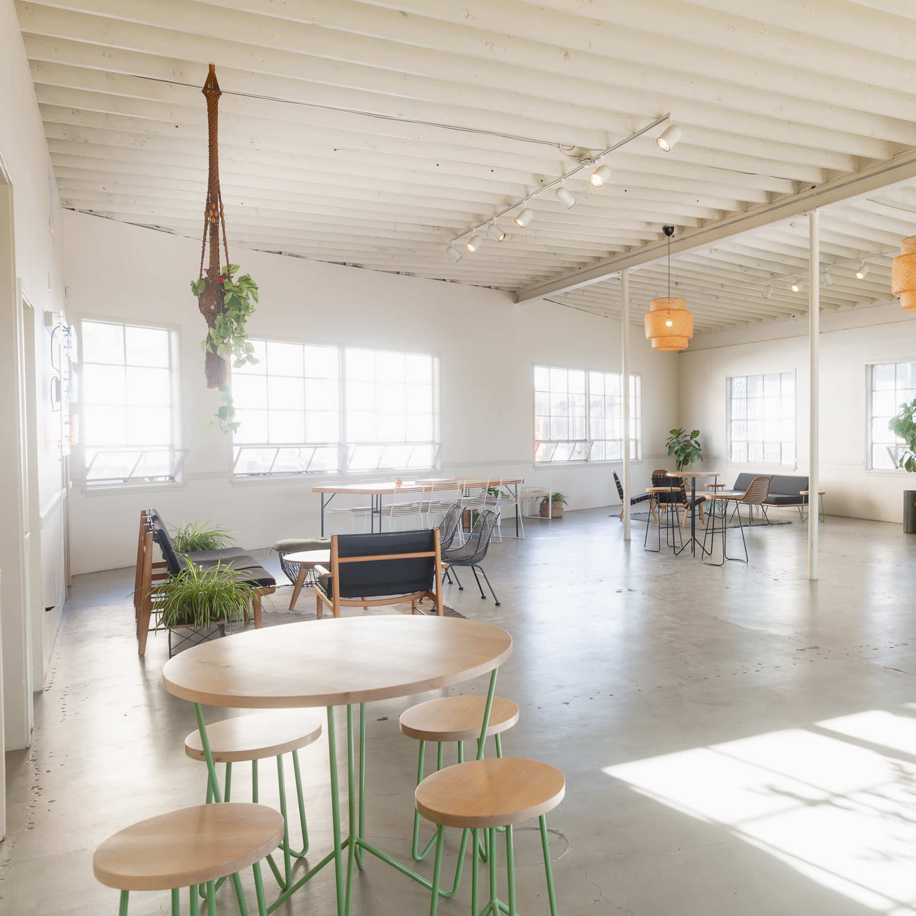 A spacious, well-lit interior with wooden beams, various tables and chairs, and large windows allowing natural light to fill the space.