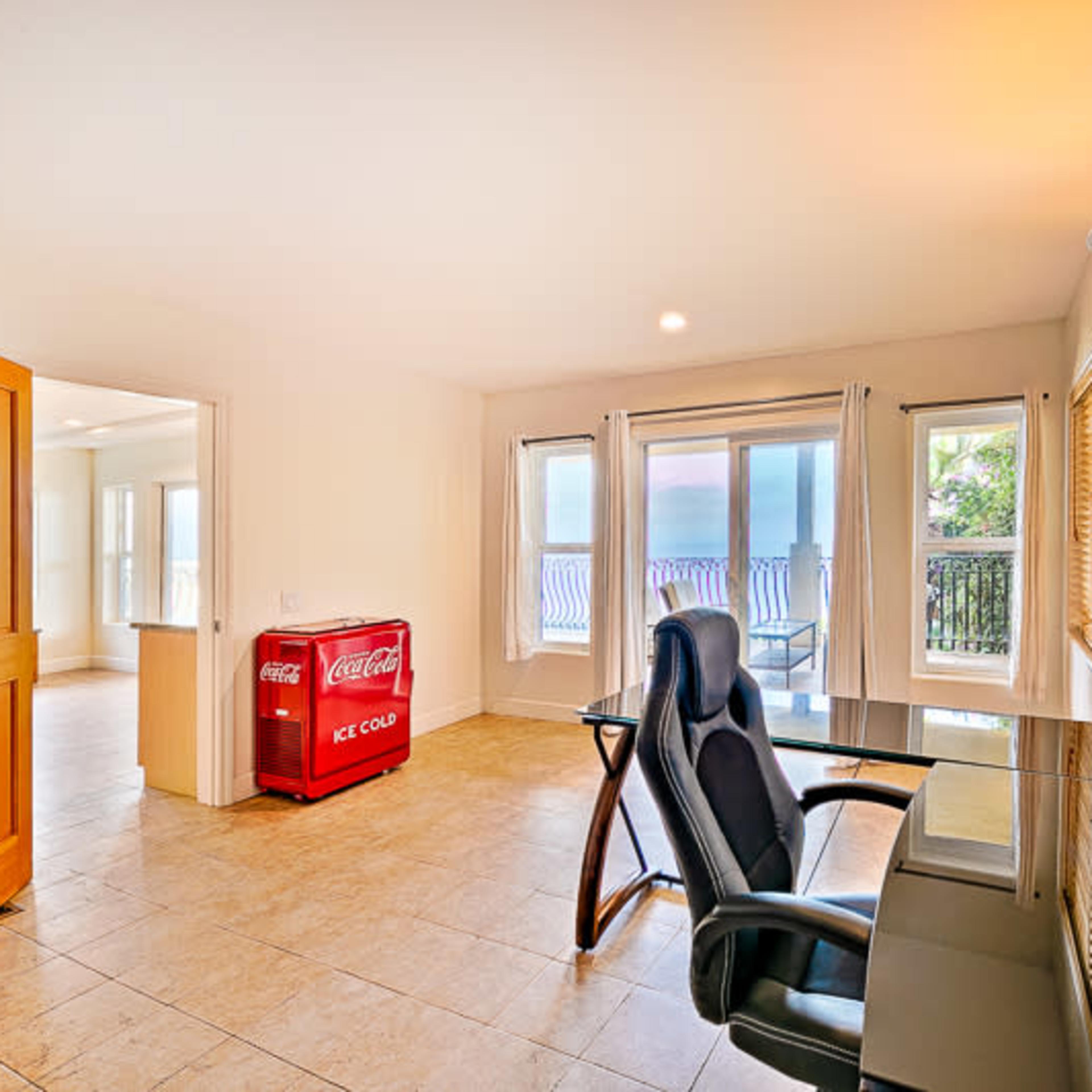 A home office features a desk and chair with a Coca-Cola ice chest in the background, positioned next to large windows that lead to a balcony.