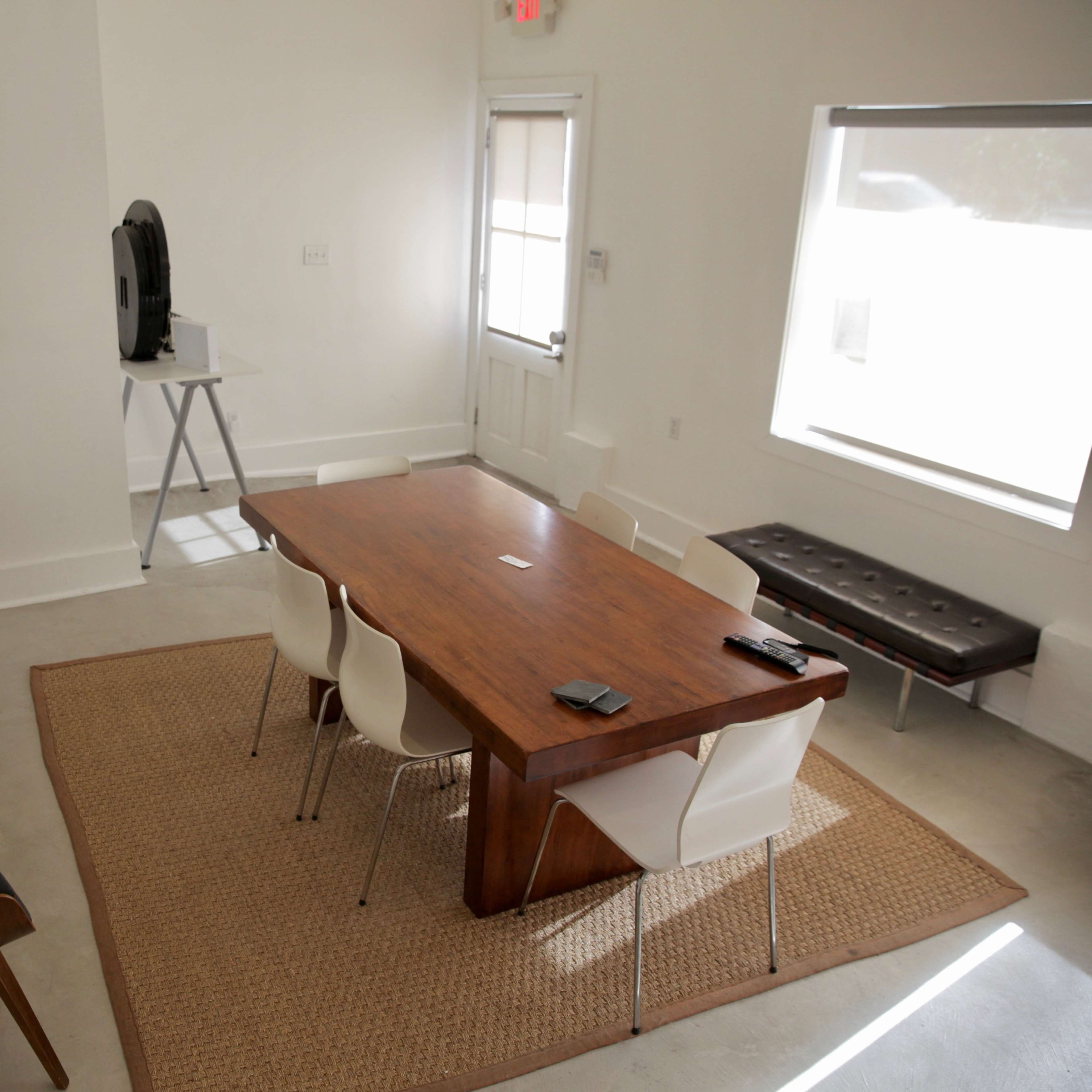 A modern conference room features a wooden table surrounded by white chairs, a large plant in the corner, and a seating area with a couch and lamp.