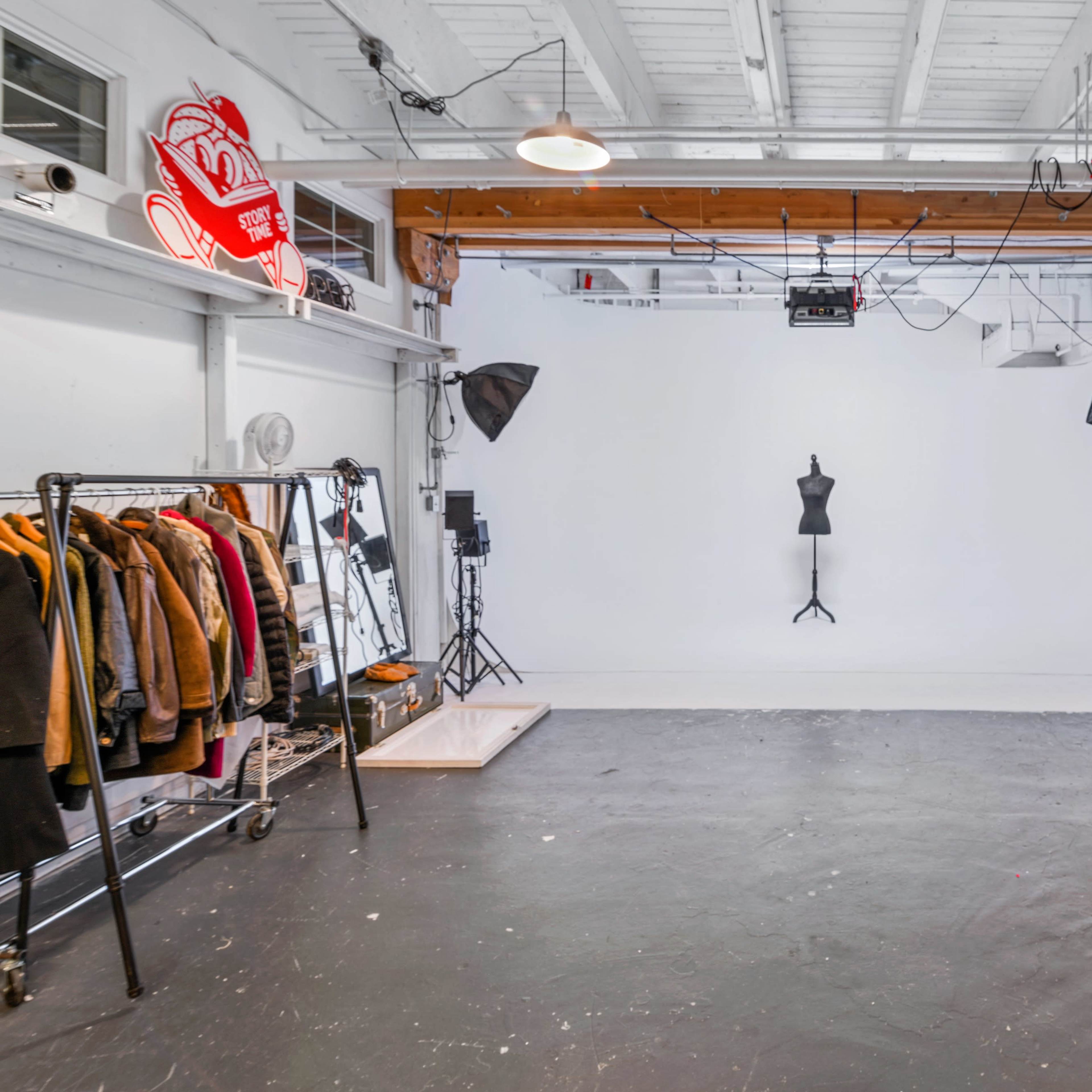 A spacious studio with a white backdrop, a clothing rack with various garments, and professional lighting equipment set up around a mannequin.