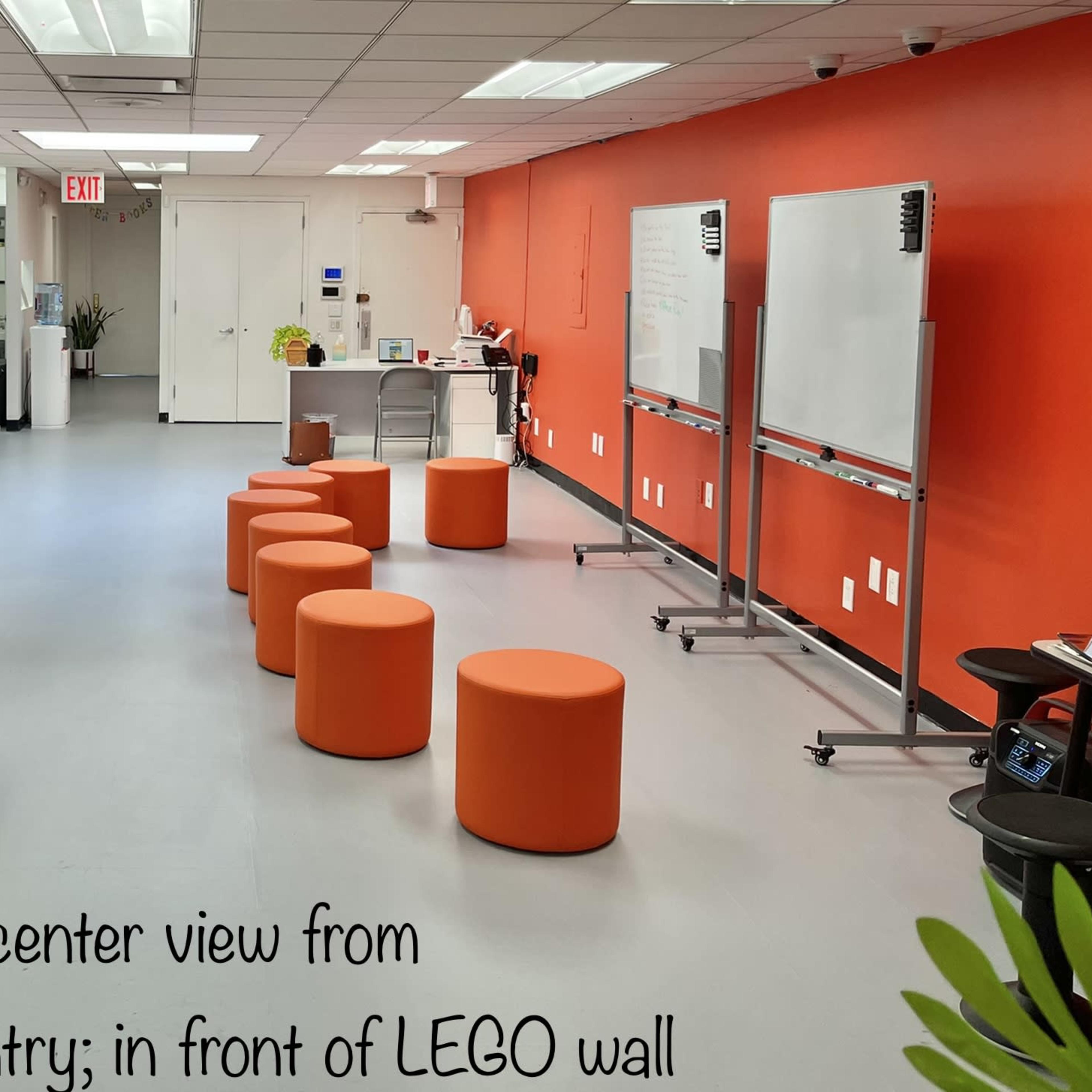 A bright room with orange walls, featuring tables and chairs on one side and orange stools arranged in the center, while a LEGO wall and whiteboards are visible on the other side.