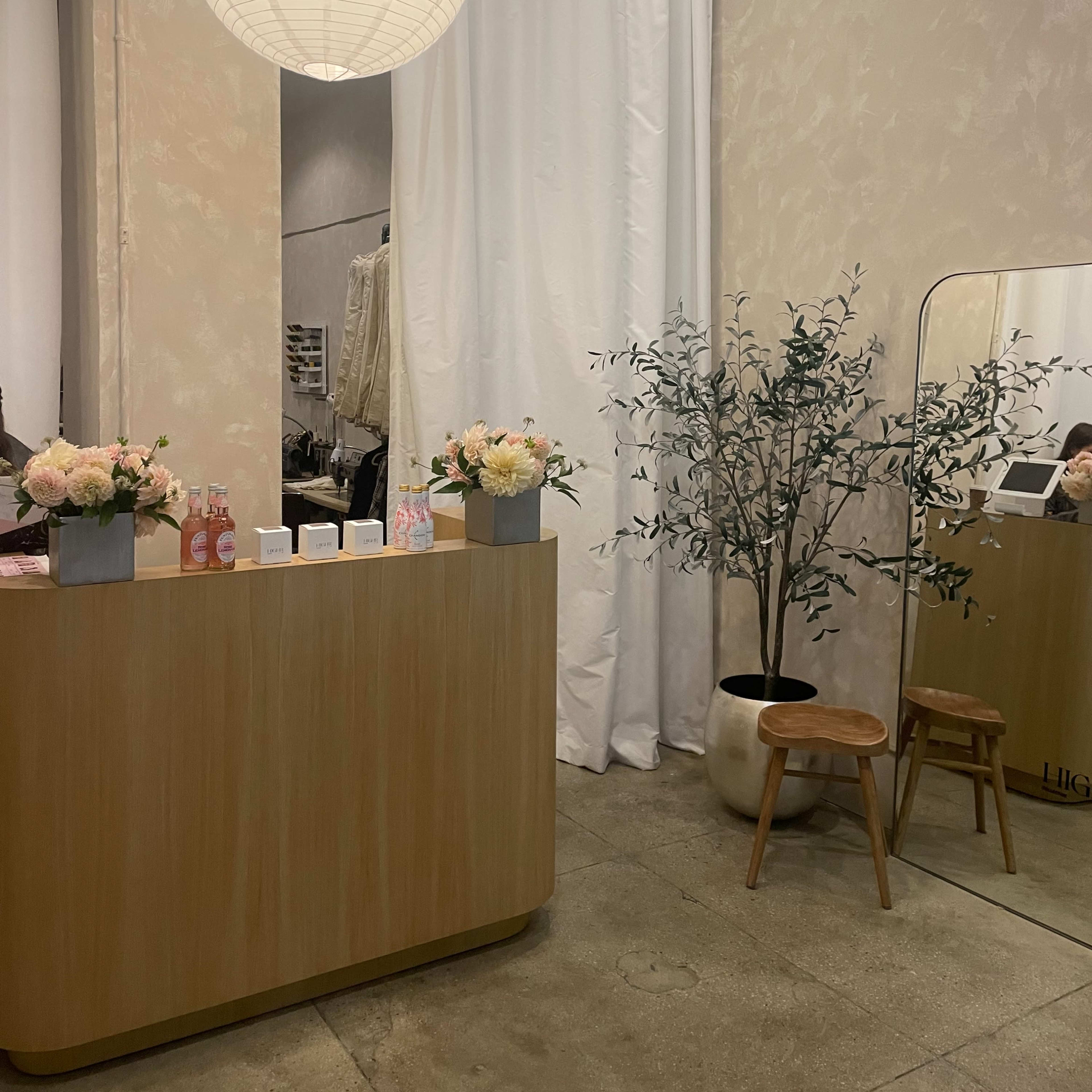A bright interior of a retail space featuring a wooden reception desk, a large round pendant light, a mirror, a potted plant, and a white curtain dividing the area.