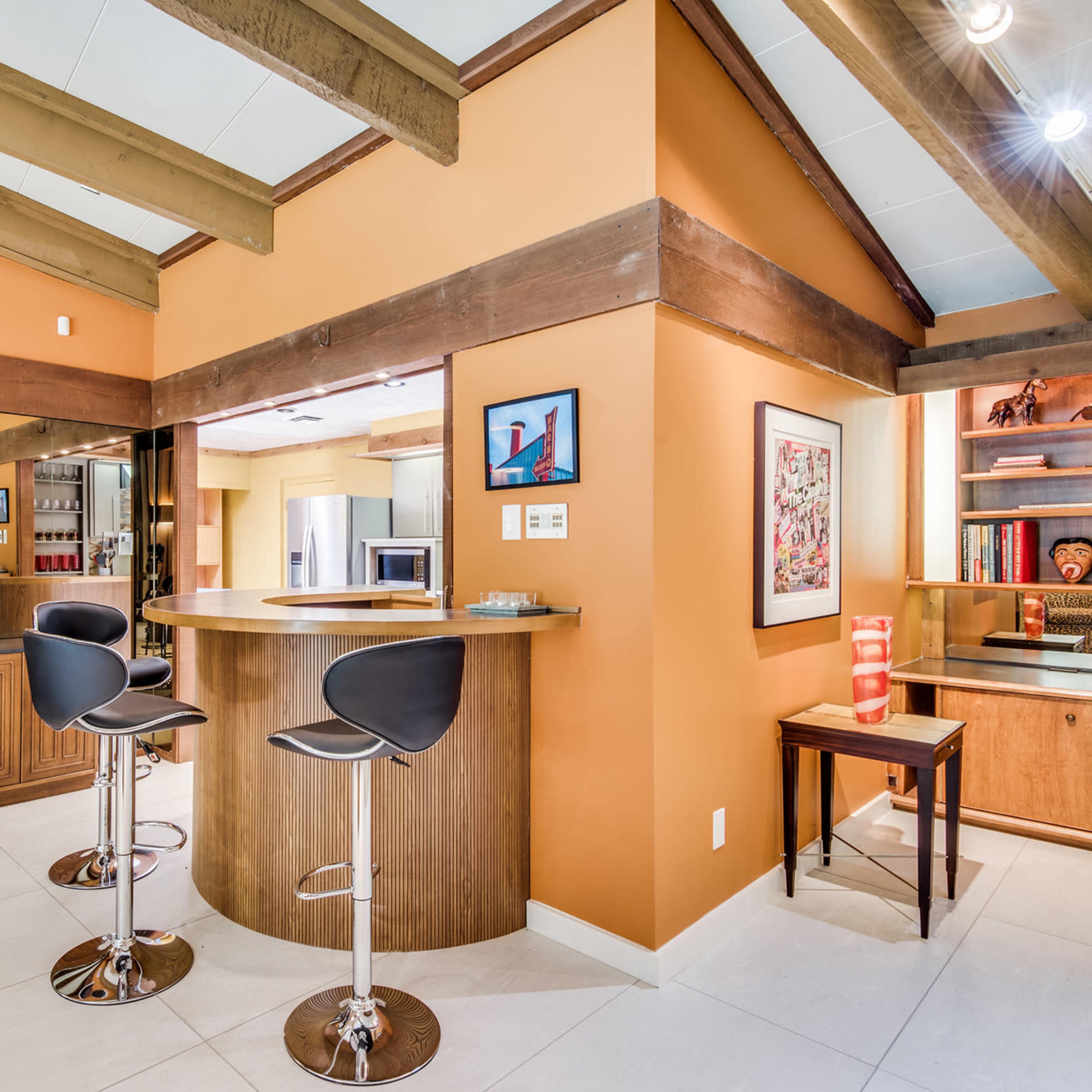 A modern interior space featuring a circular bar with two high stools, warm orange walls, wooden beams, and a blend of contemporary and decorative elements.