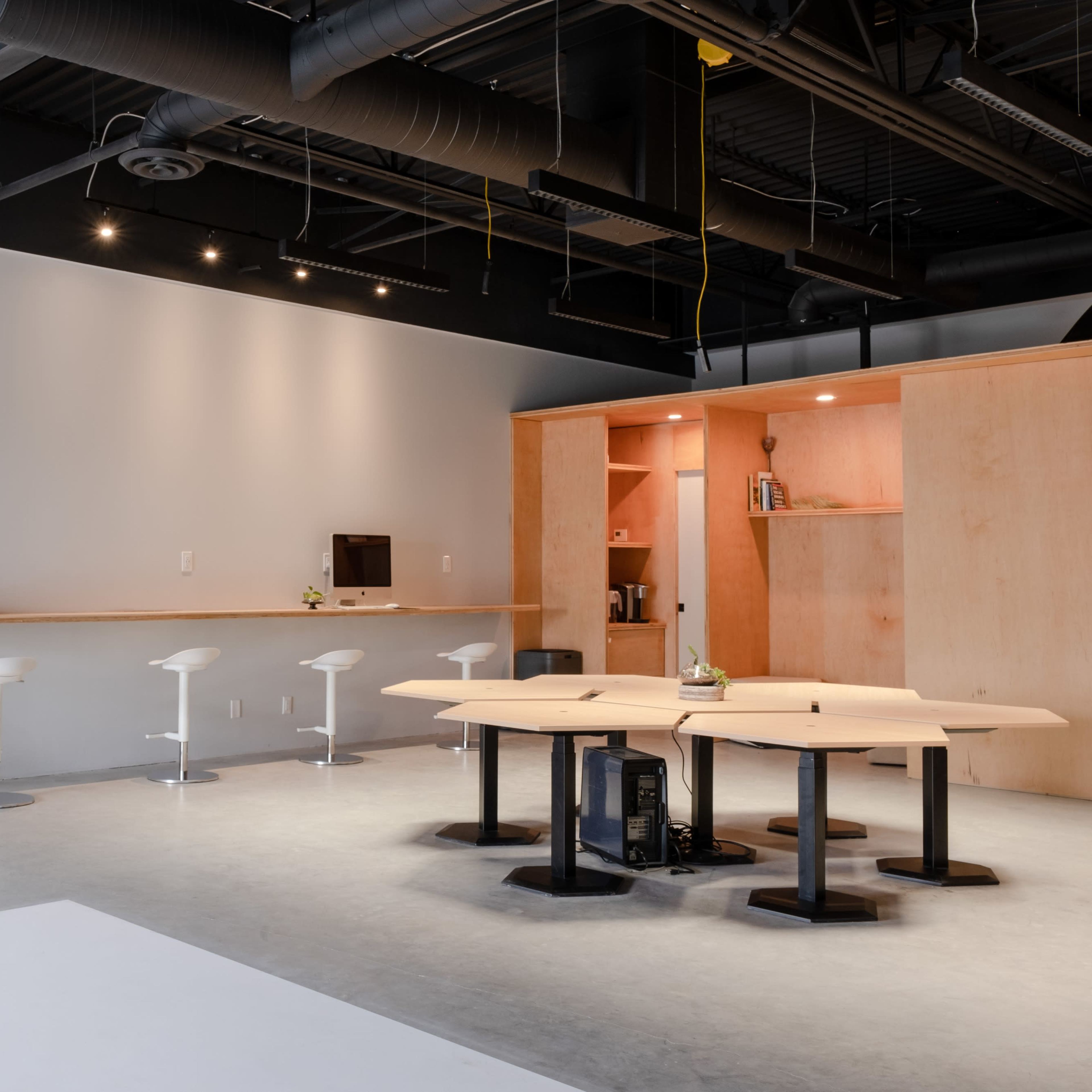 A modern open office space featuring a wooden meeting table, white bar stools, and minimalistic cabinetry against a light gray wall.