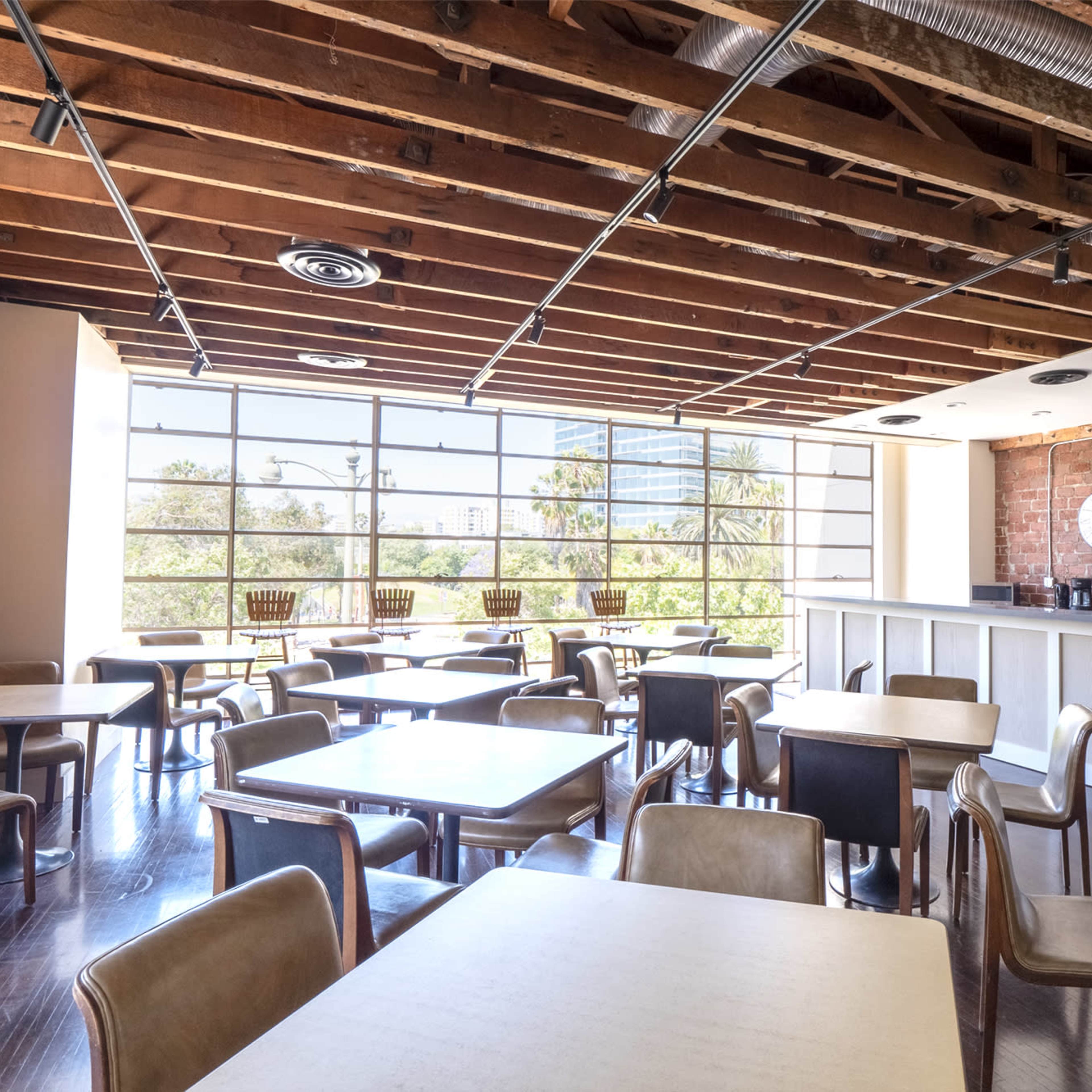 A cafe with wooden beams overhead, brick walls, and numerous empty tables arranged for dining next to large windows.