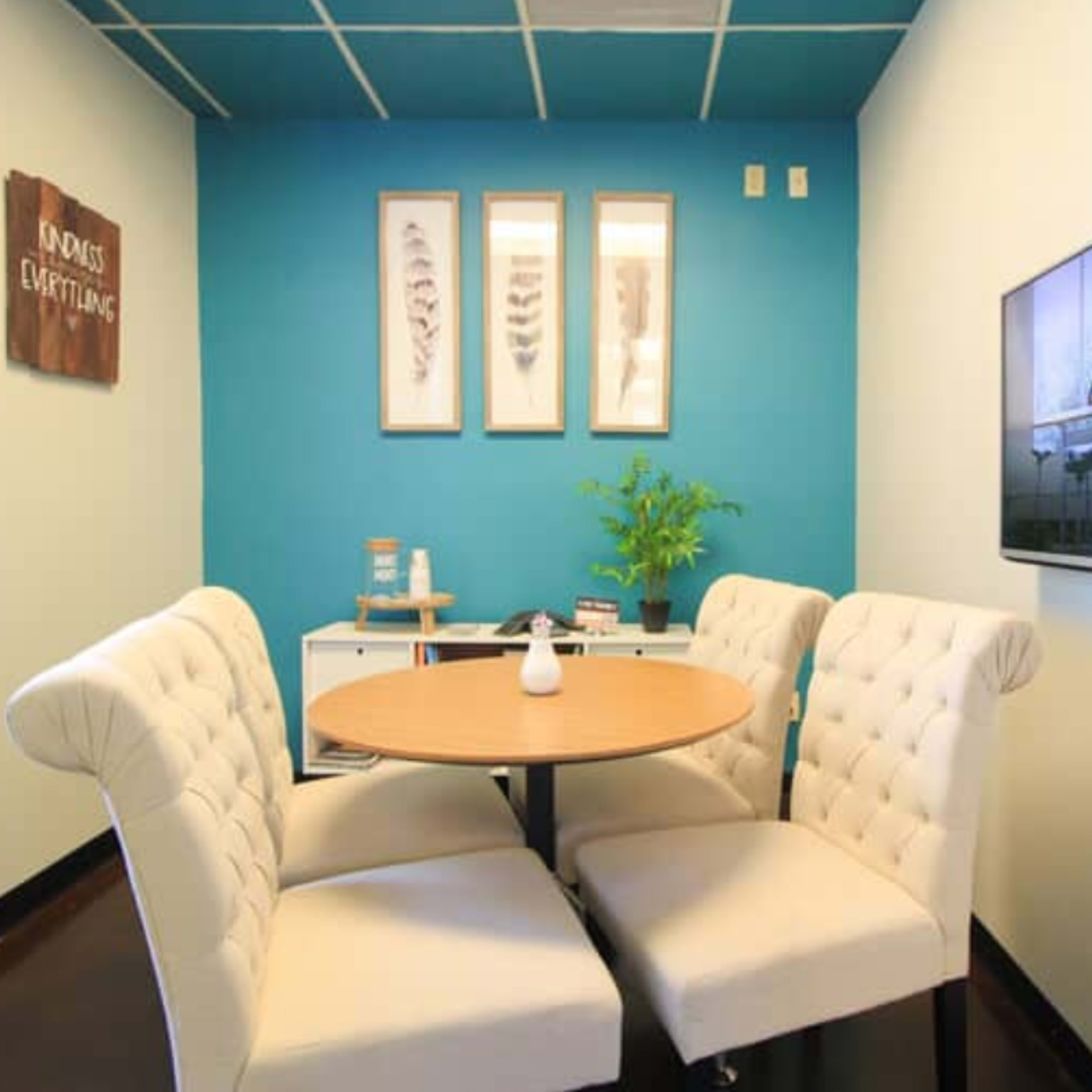 A small meeting area features a round table with four tufted chairs and a television mounted on the wall, alongside a plant and framed wall art.