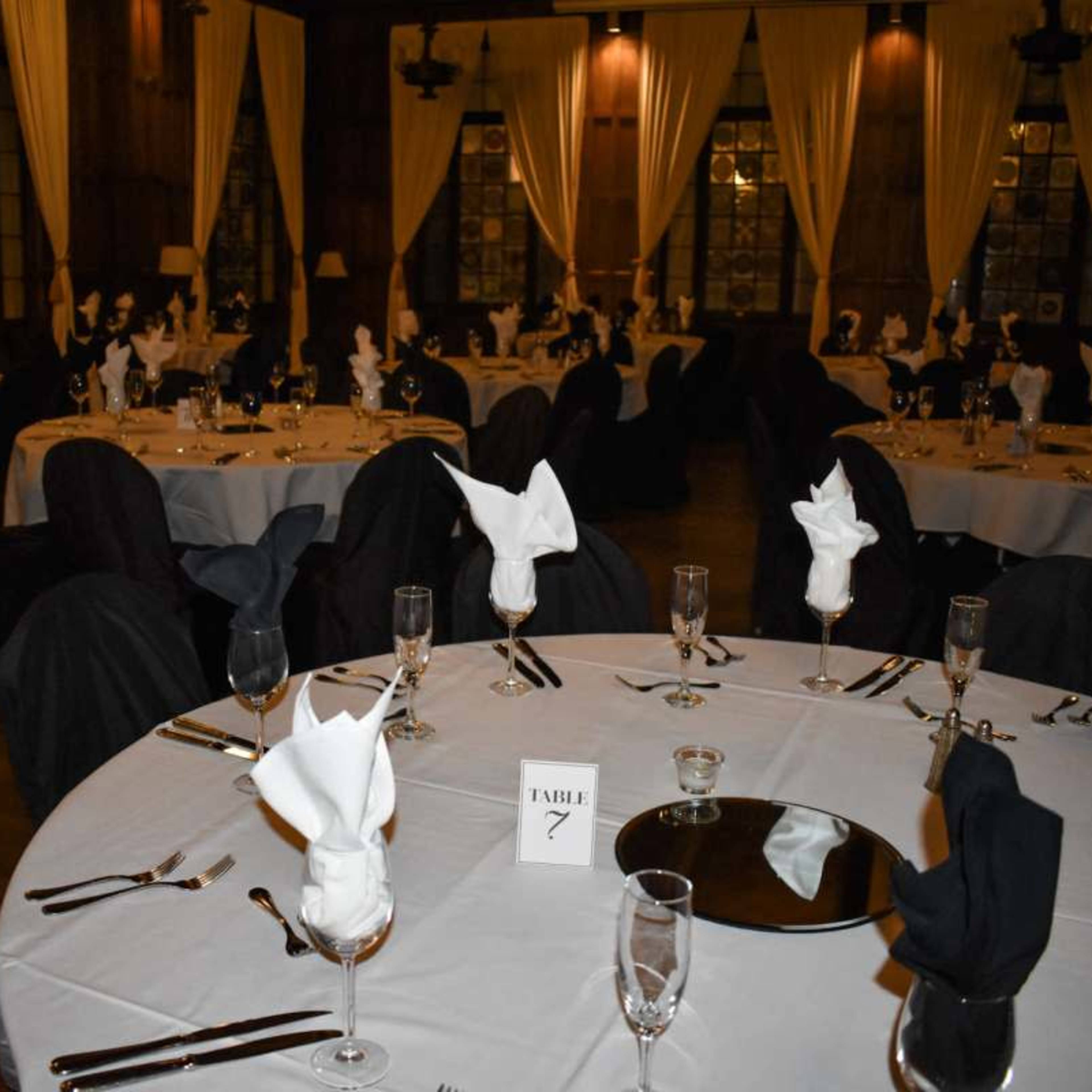A formal dining room is set with multiple round tables covered in white tablecloths, featuring neatly arranged glassware and folded napkins in black and white.