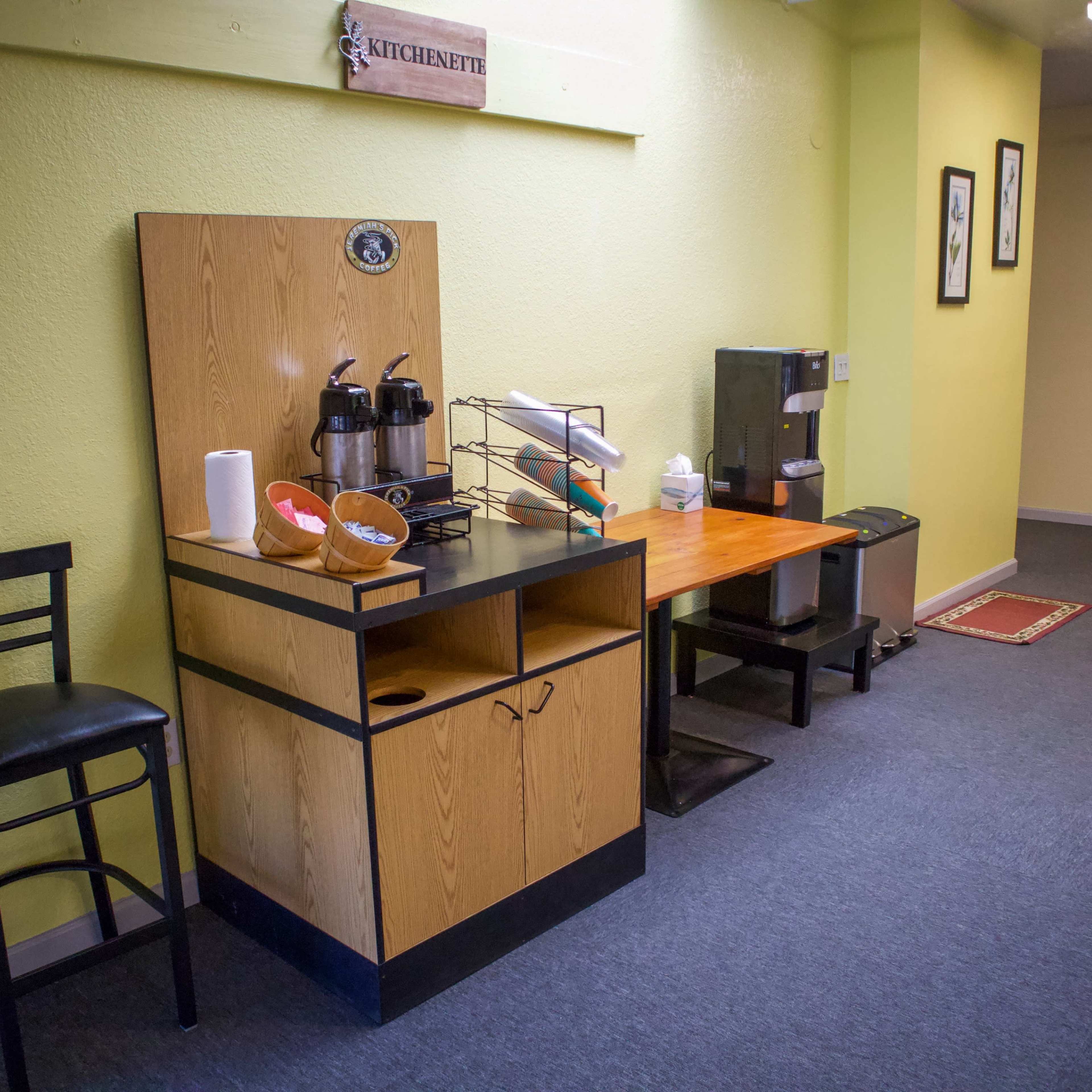 A kitchenette area features a wooden counter with coffee dispensers, a roll of paper towels, and a small black countertop appliance, beside a chair and framed pictures on the green walls.