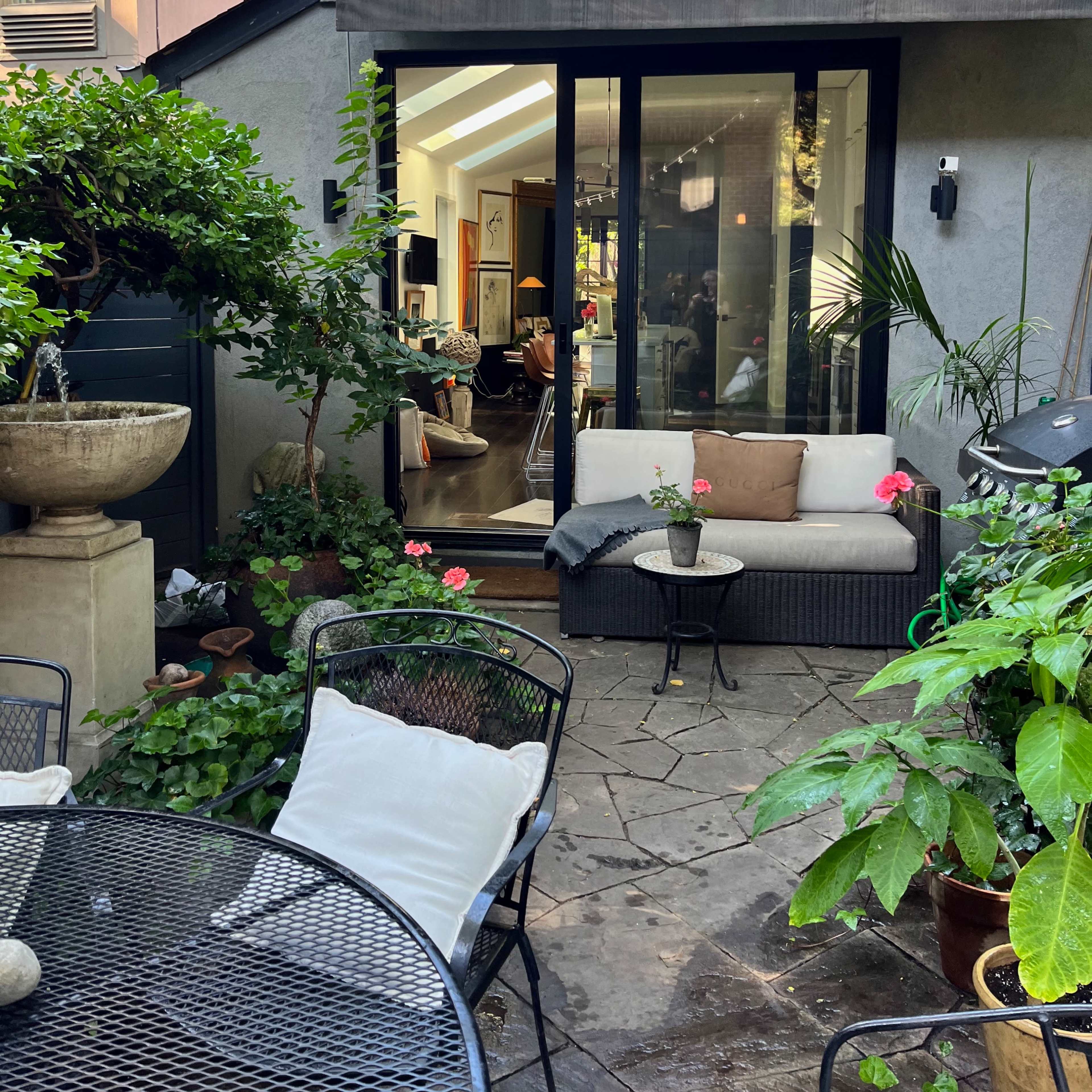 A cozy outdoor seating area with stone flooring, surrounded by plants and featuring a view of a modern interior through a large glass door.