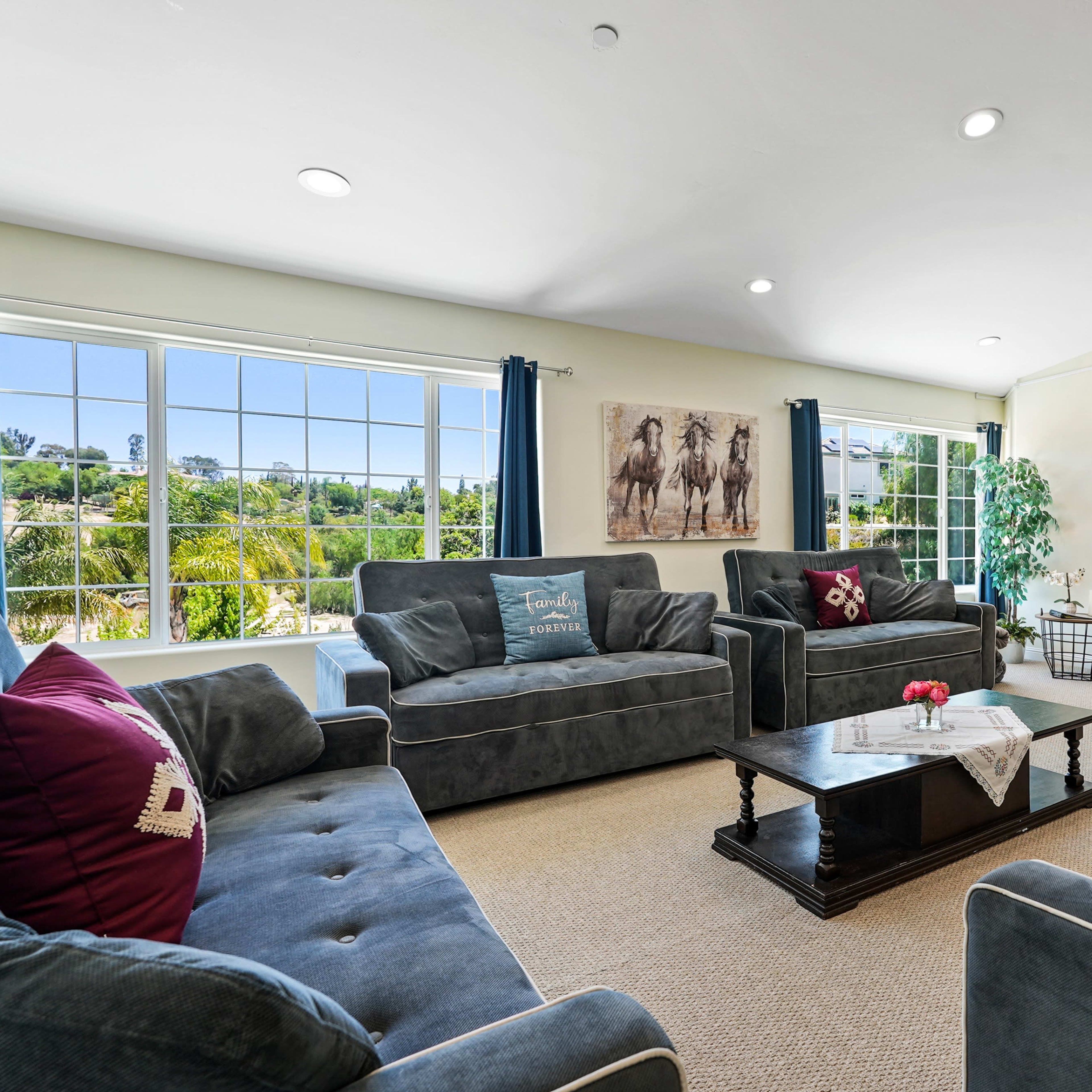 The living room features gray sofas arranged around a central table, with large windows providing a view of greenery outside and a wall-mounted television.