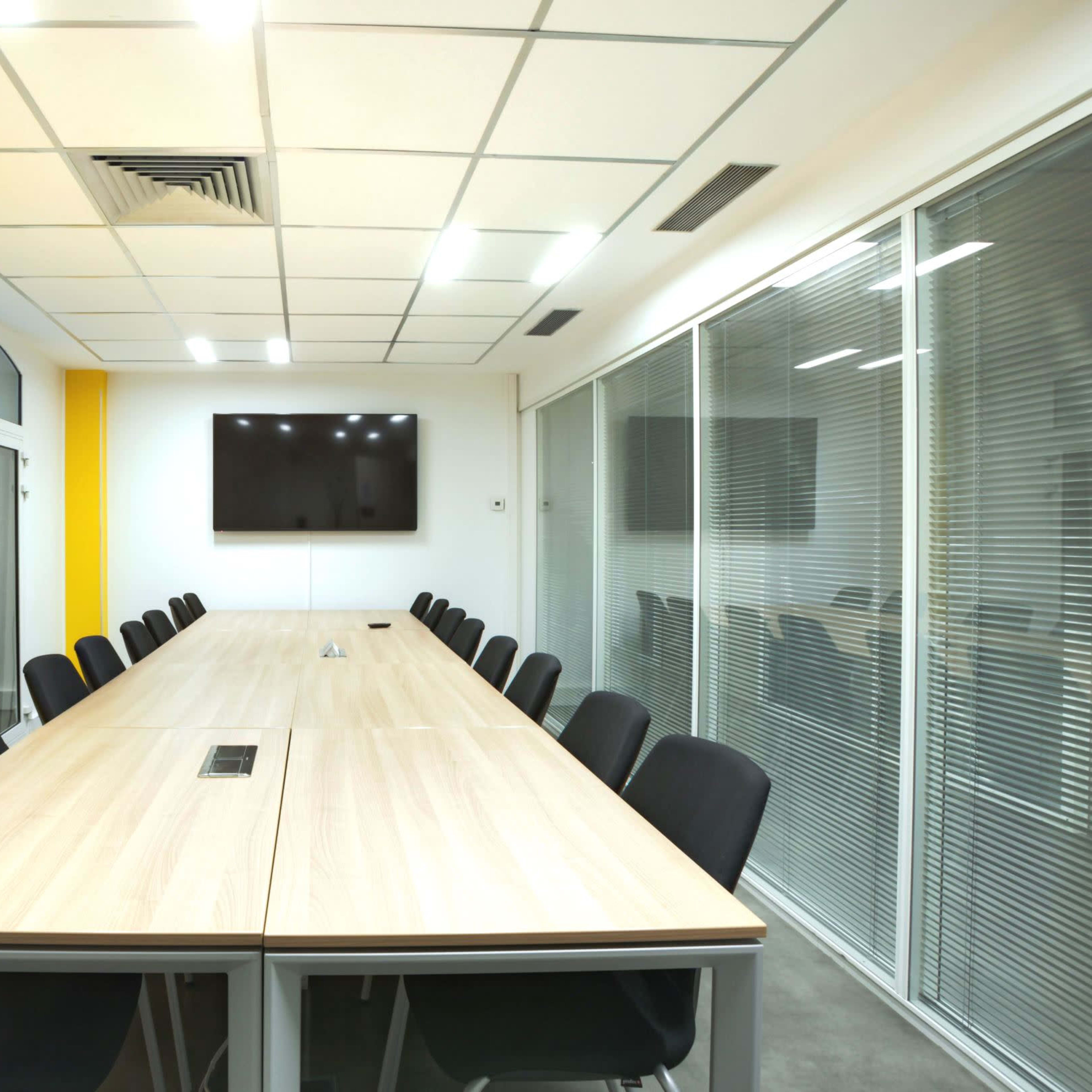 A modern conference room features a long wooden table surrounded by black chairs, with a large television mounted on the wall and a bright yellow accent wall.