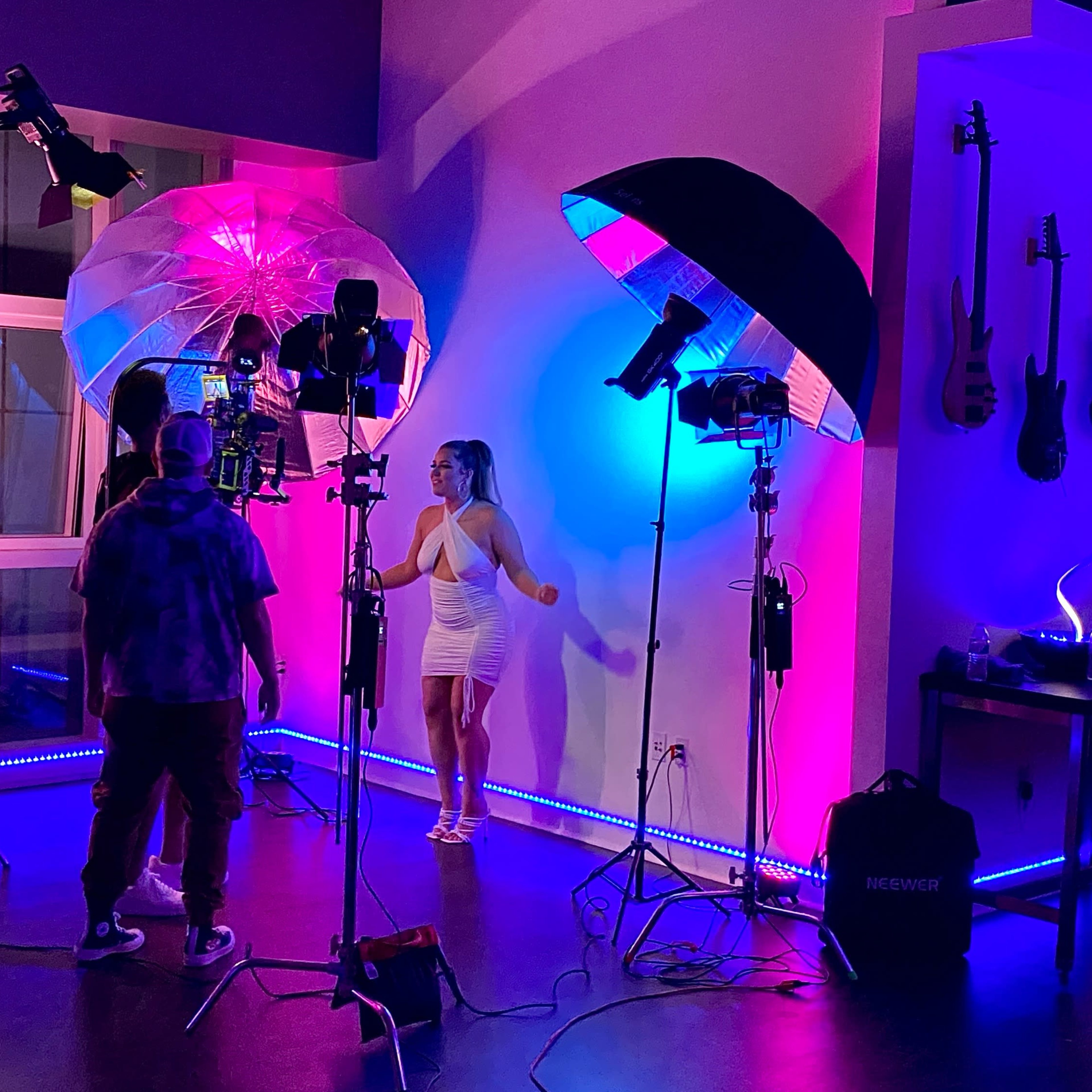 A photoshoot takes place in a studio with colorful lights, equipment, and a model posing in a white dress.