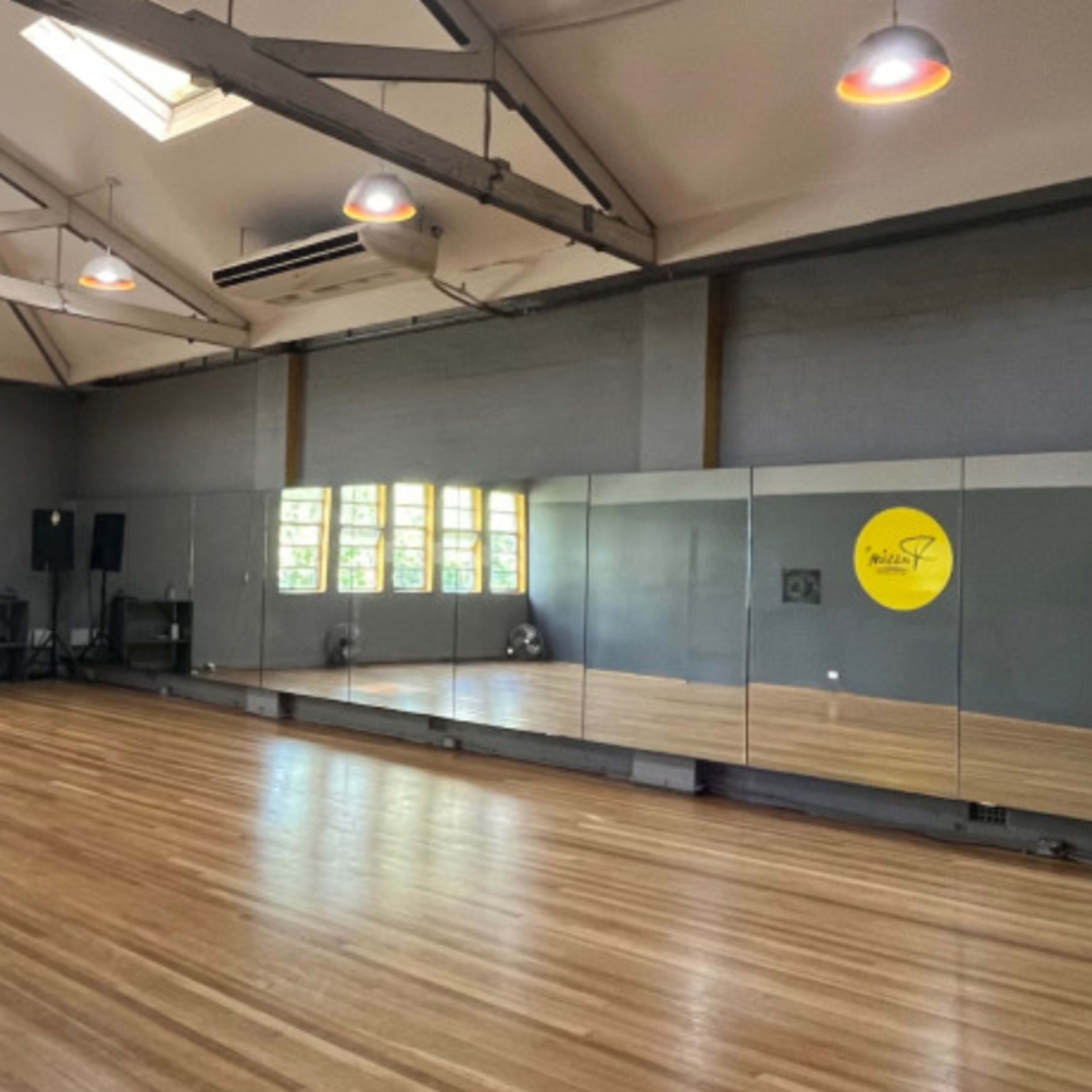 A spacious dance studio features a wooden floor, large mirrors along one wall, and several windows allowing natural light.