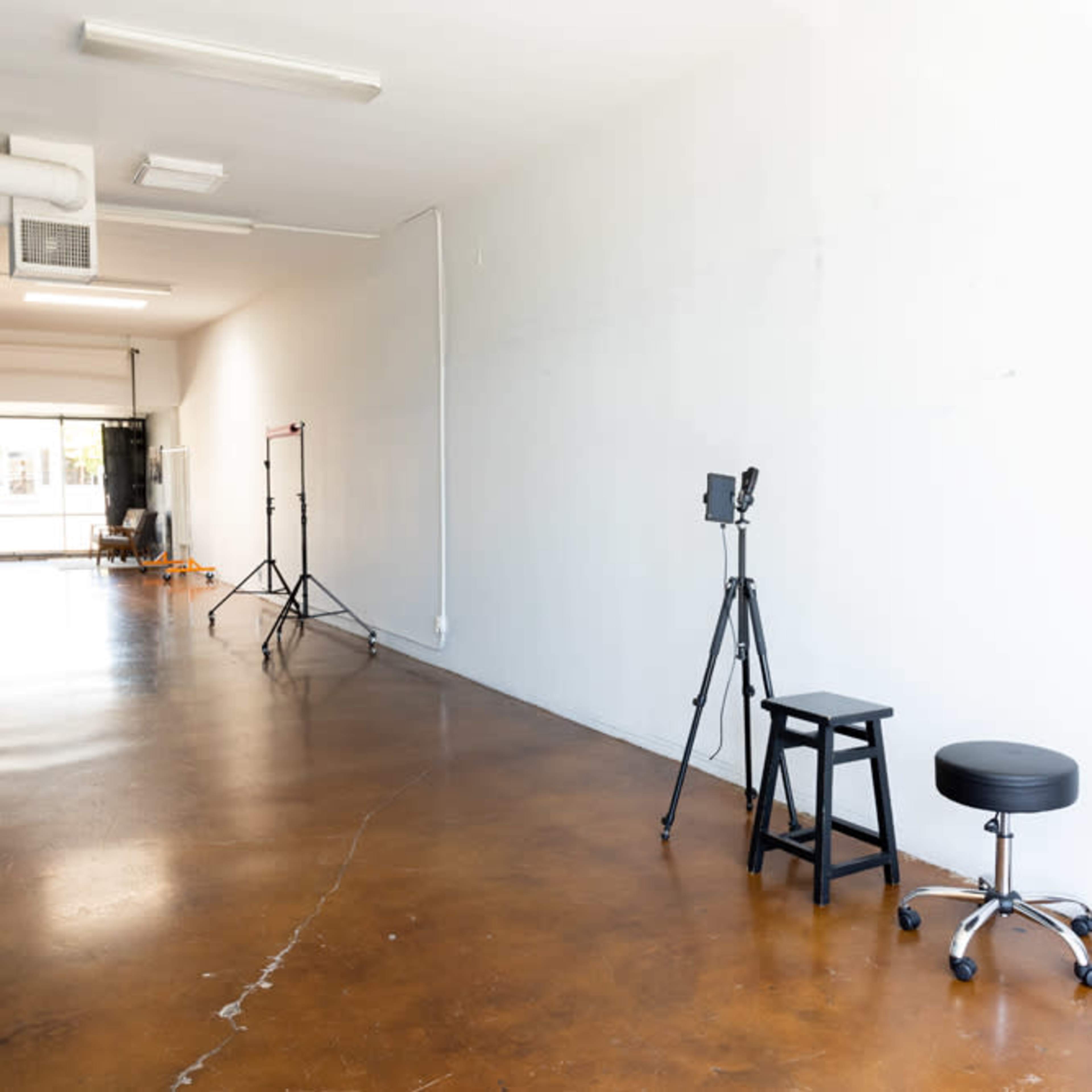 A spacious, well-lit studio features two tripods, a stool, and a black rolling chair along a polished concrete floor.