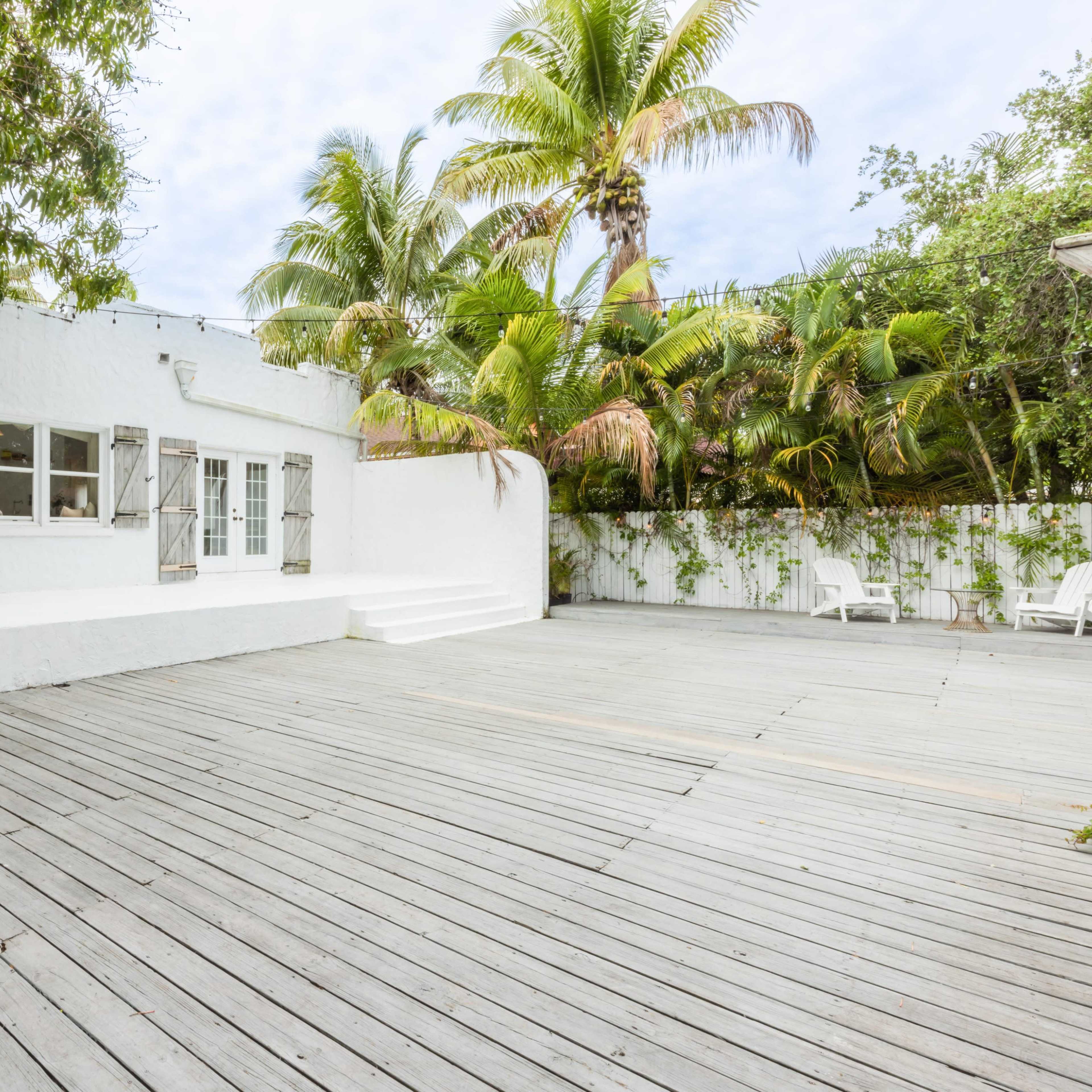 A white stucco house with wooden shutters surrounded by palm trees and a spacious wooden deck.