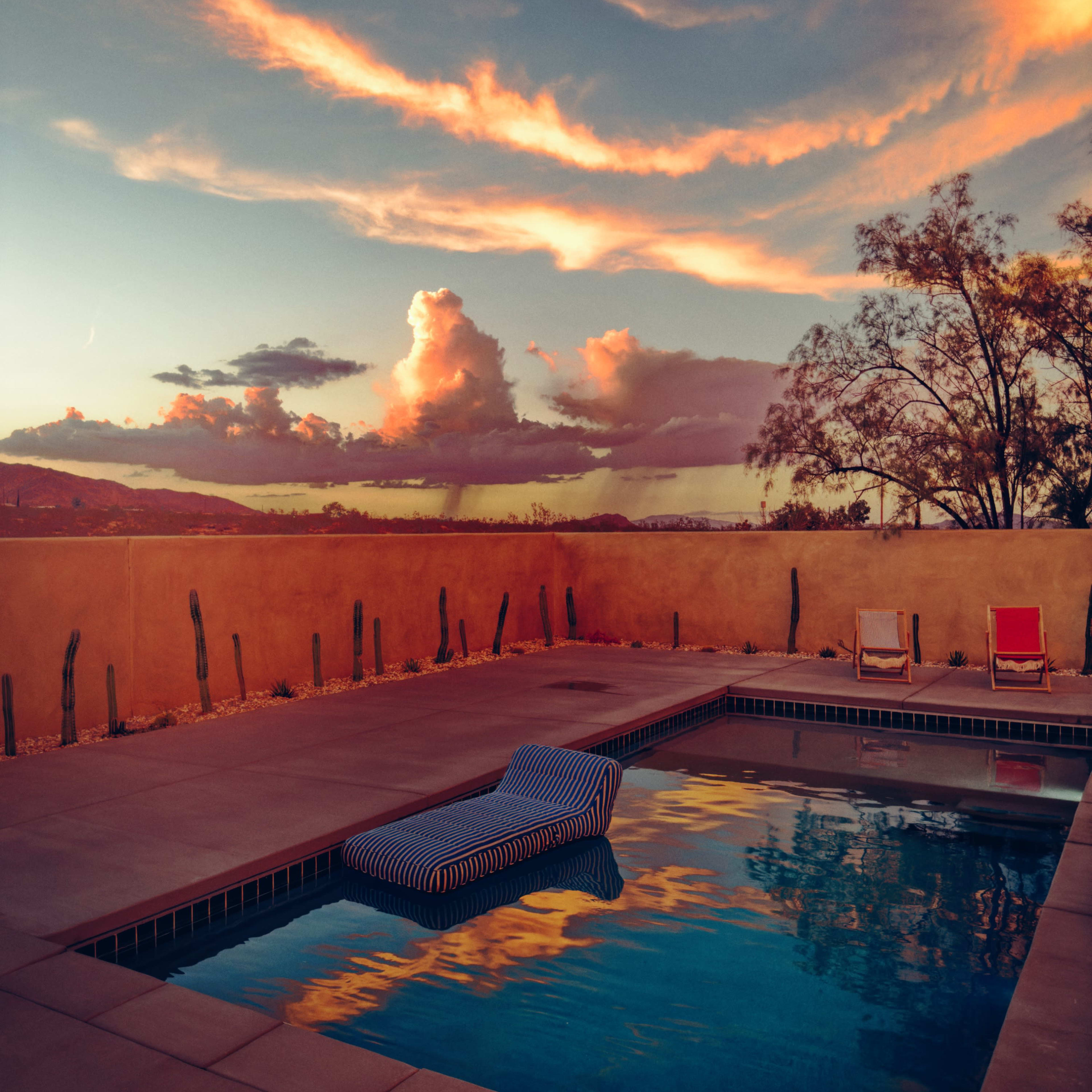 A striped lounge chair rests by a swimming pool, reflecting colorful clouds and a dusky sky.