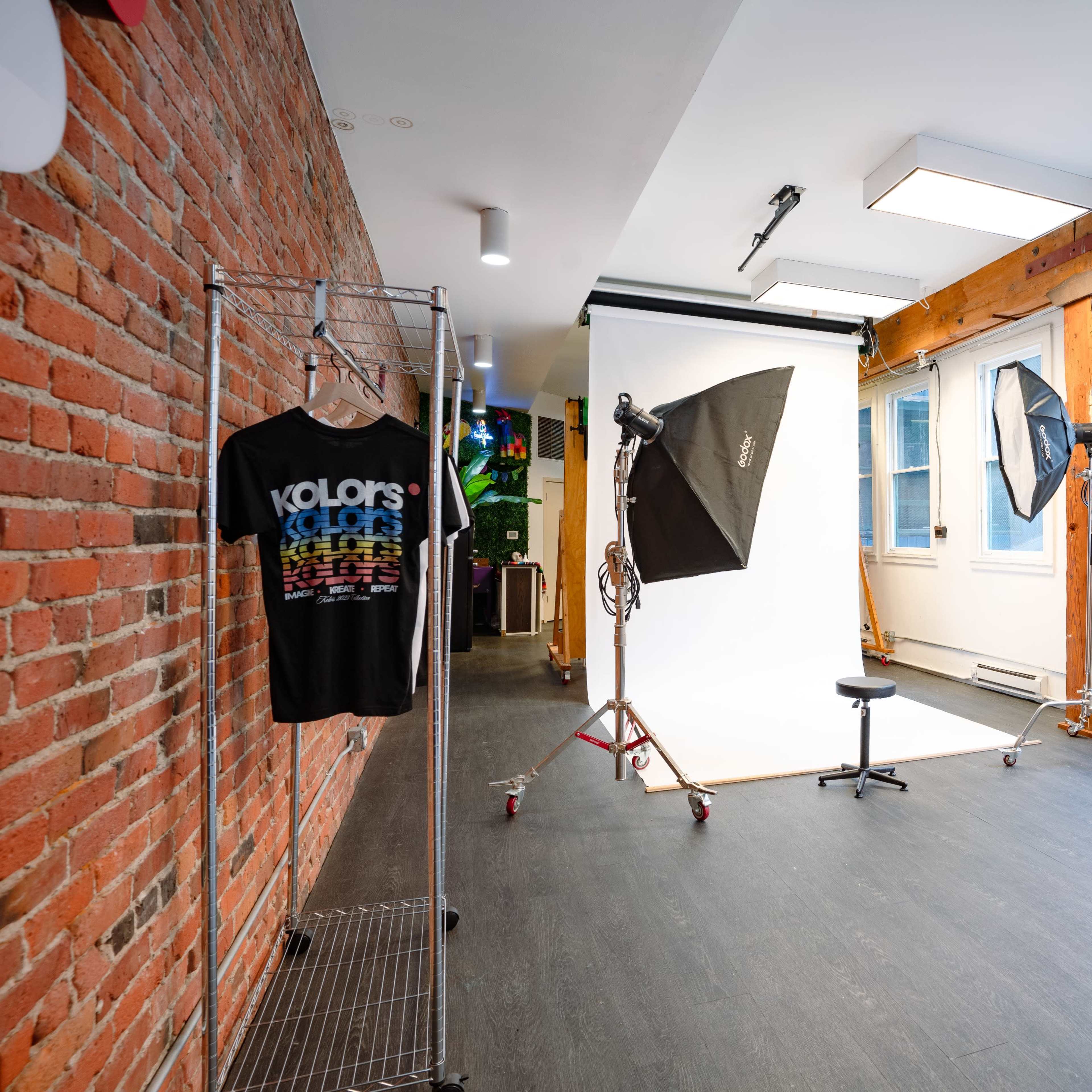 A photography studio with a white backdrop, lighting equipment, clothing rack, and a blue sofa, featuring exposed brick walls and wooden beams.