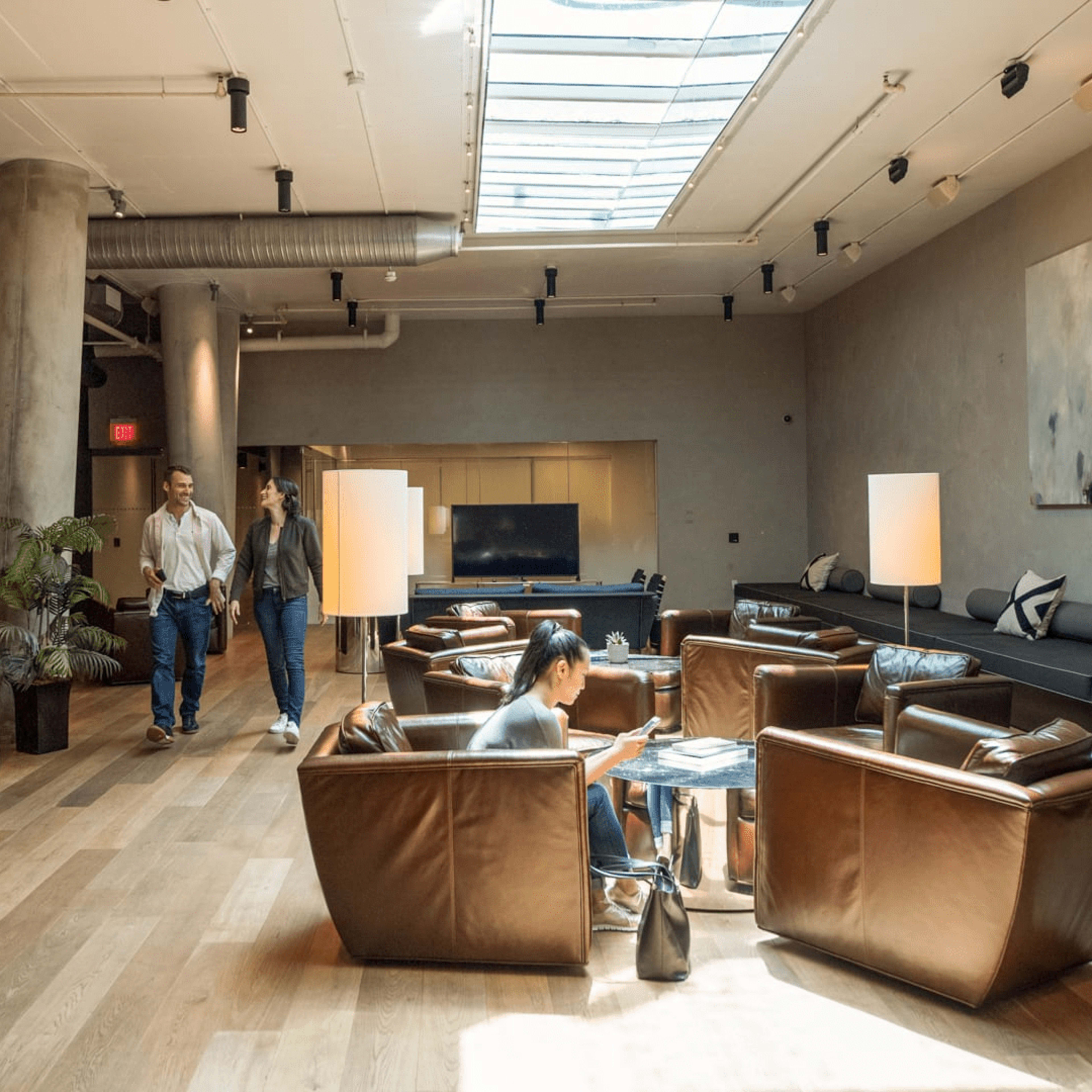 A modern lounge features leather chairs, a skylight, and two people walking through the space while another person sits at a table.