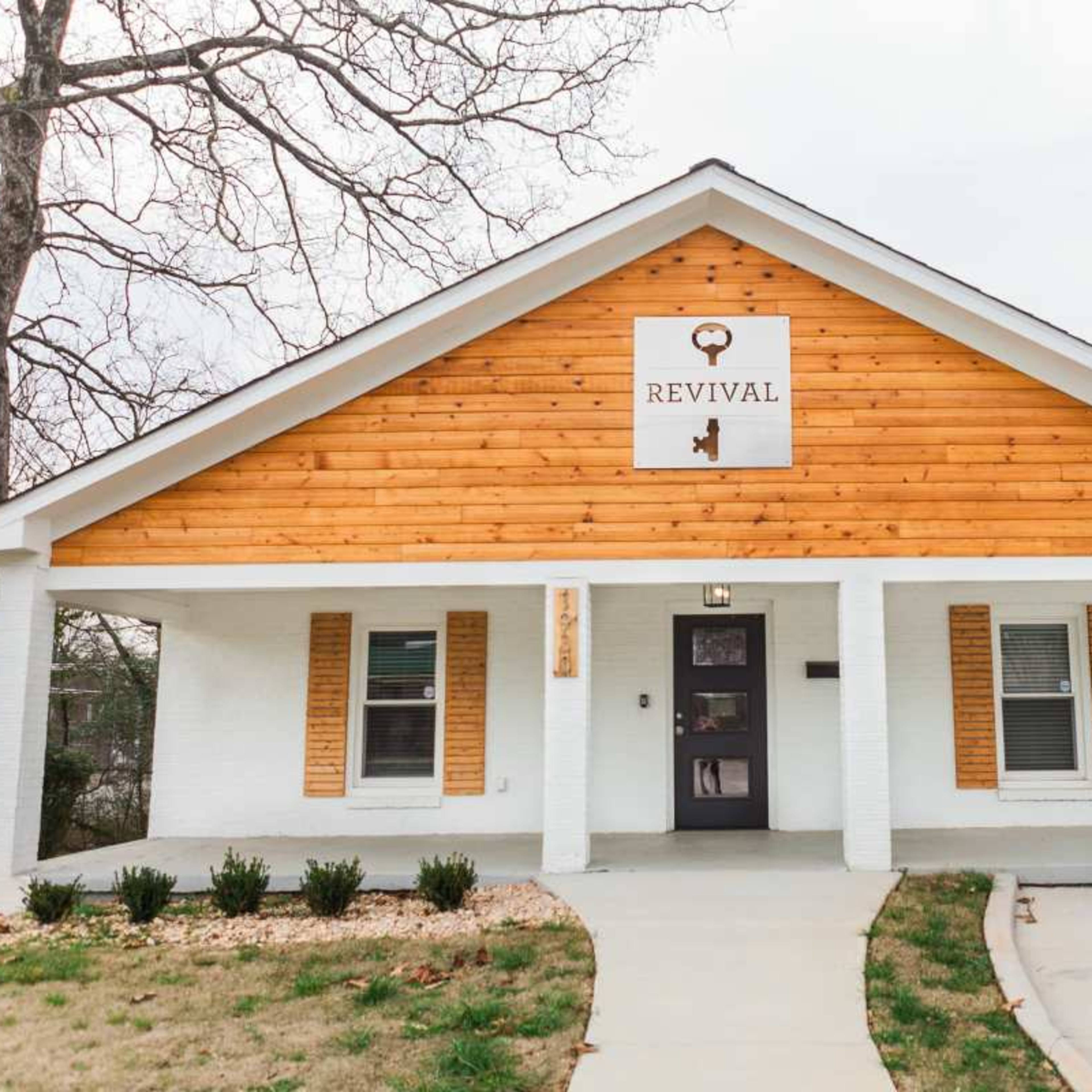 A white and wooden house features a sign labeled "REVIVAL" above the entrance, with a landscaped front yard and a paved walkway.
