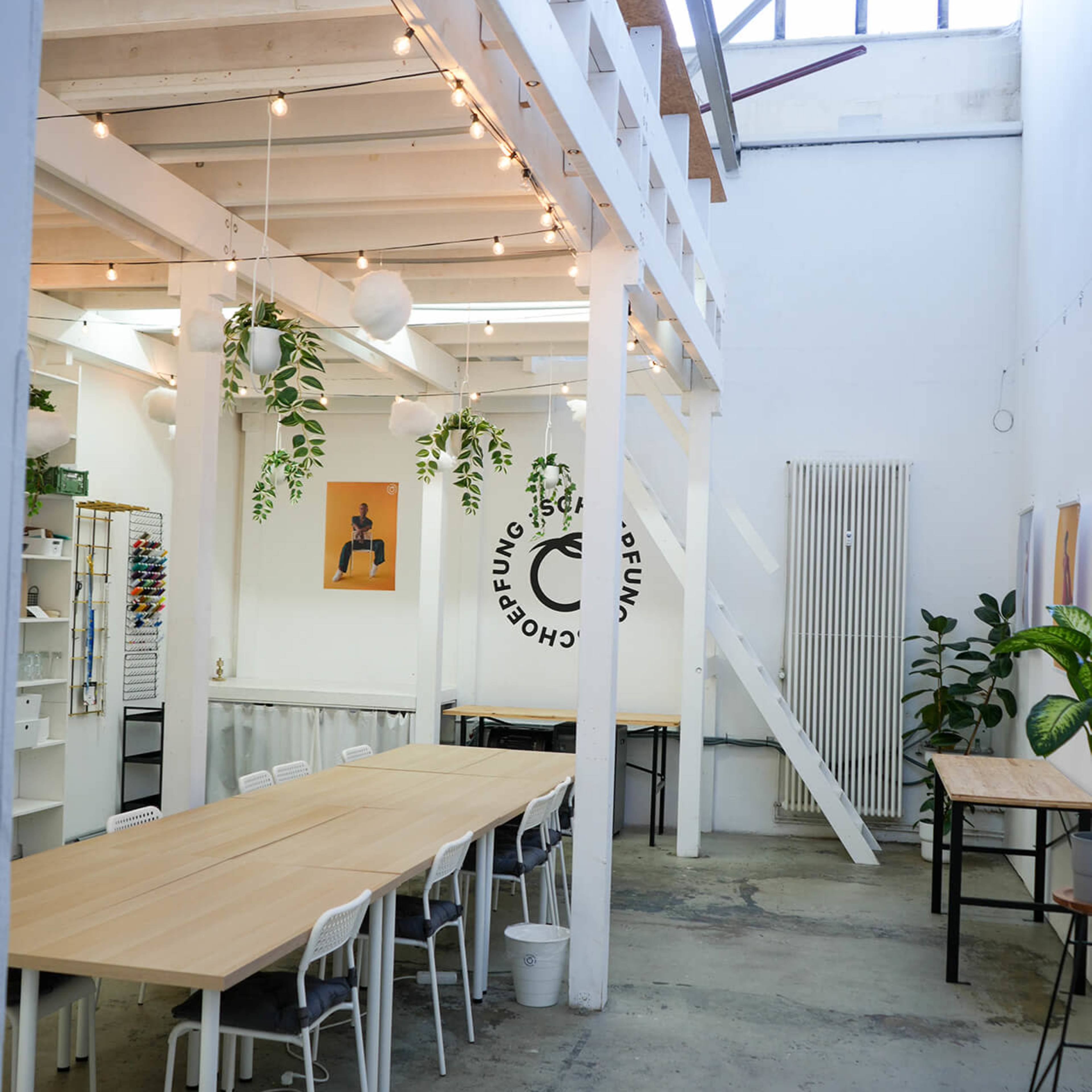 A spacious, well-lit interior with wooden tables, white walls, and plants hanging from the ceiling.