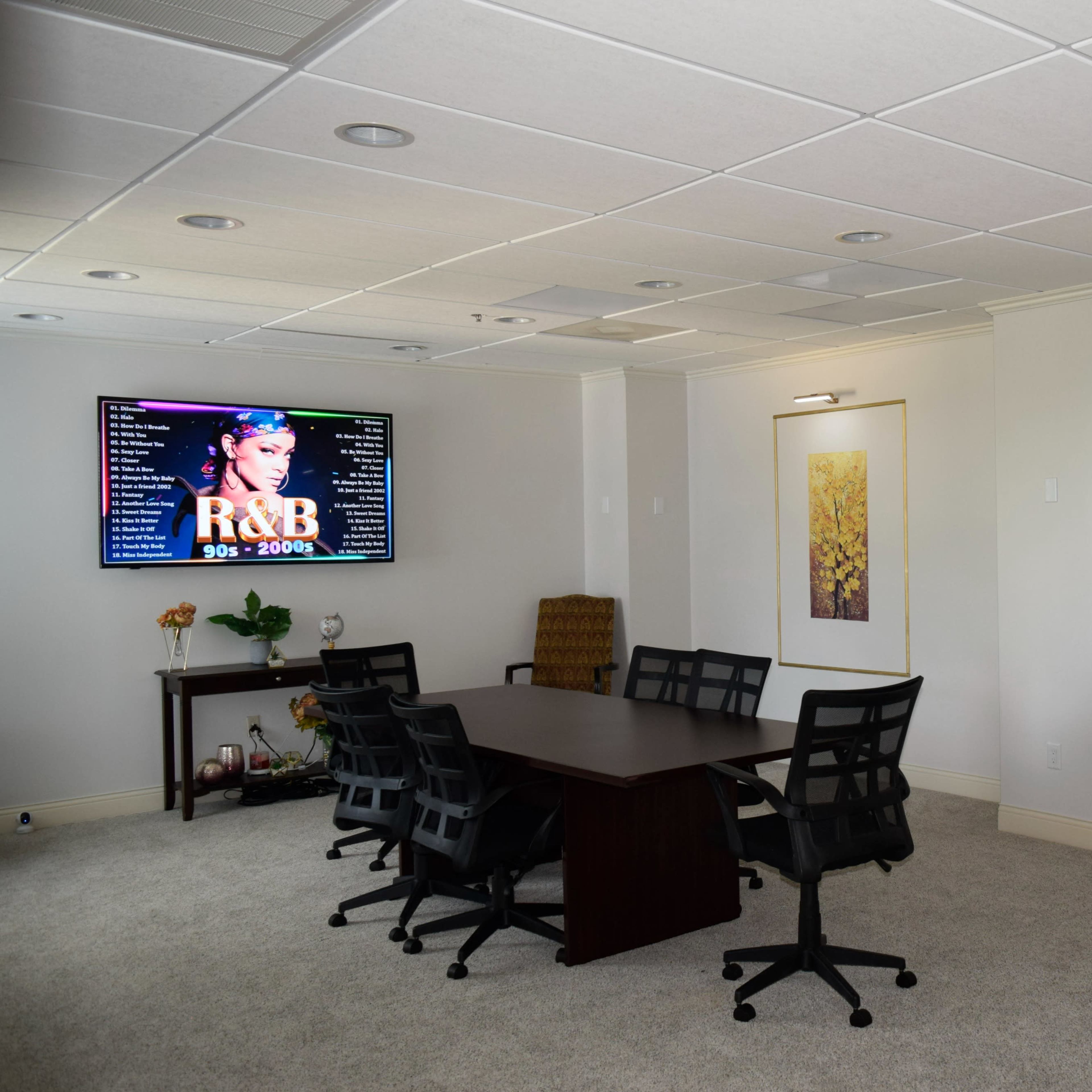 A conference room features a large screen displaying R&B music, surrounded by two framed leaf prints and a table with six chairs.