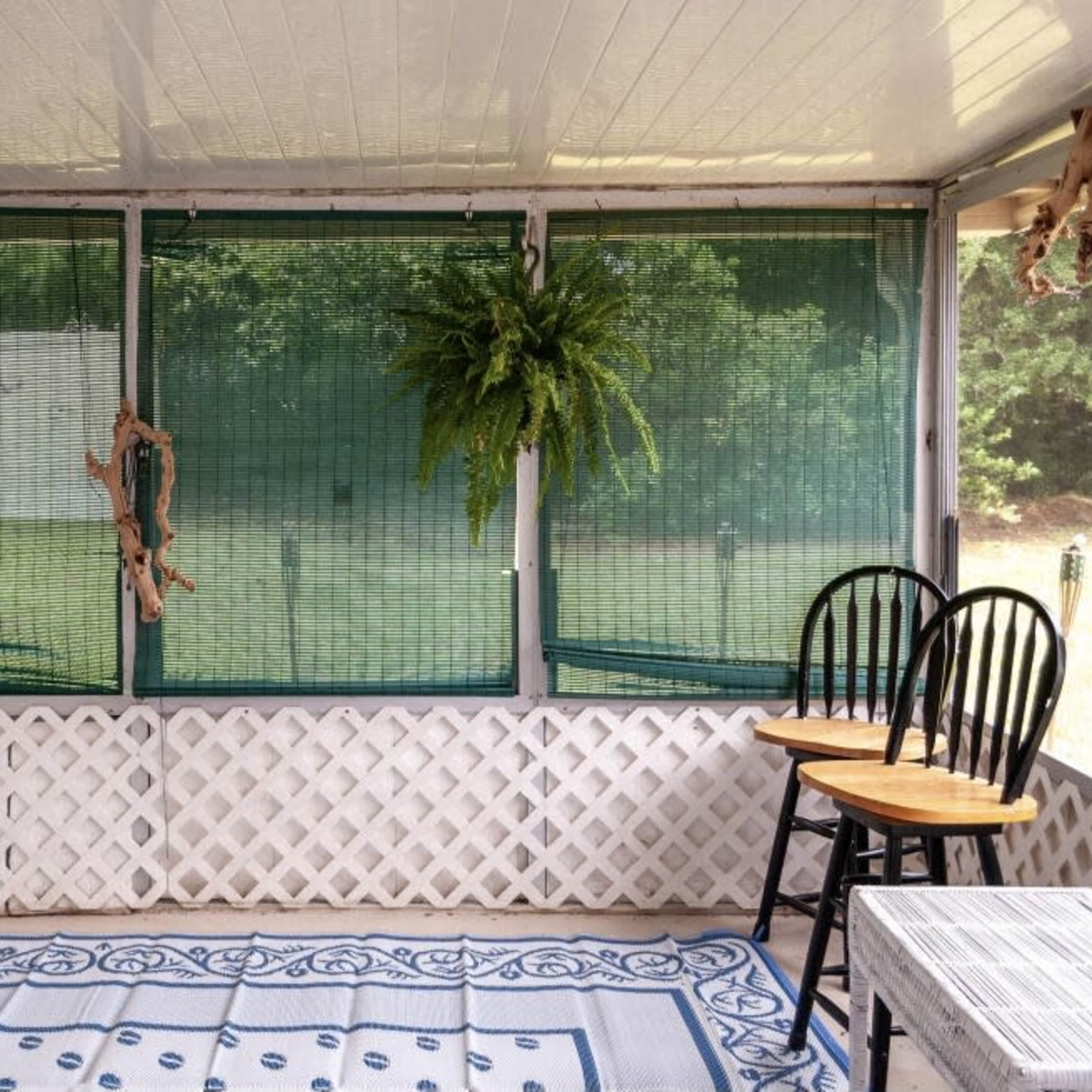 A screened porch with green window coverings, hanging ferns, and two wooden chairs beside a patterned rug.