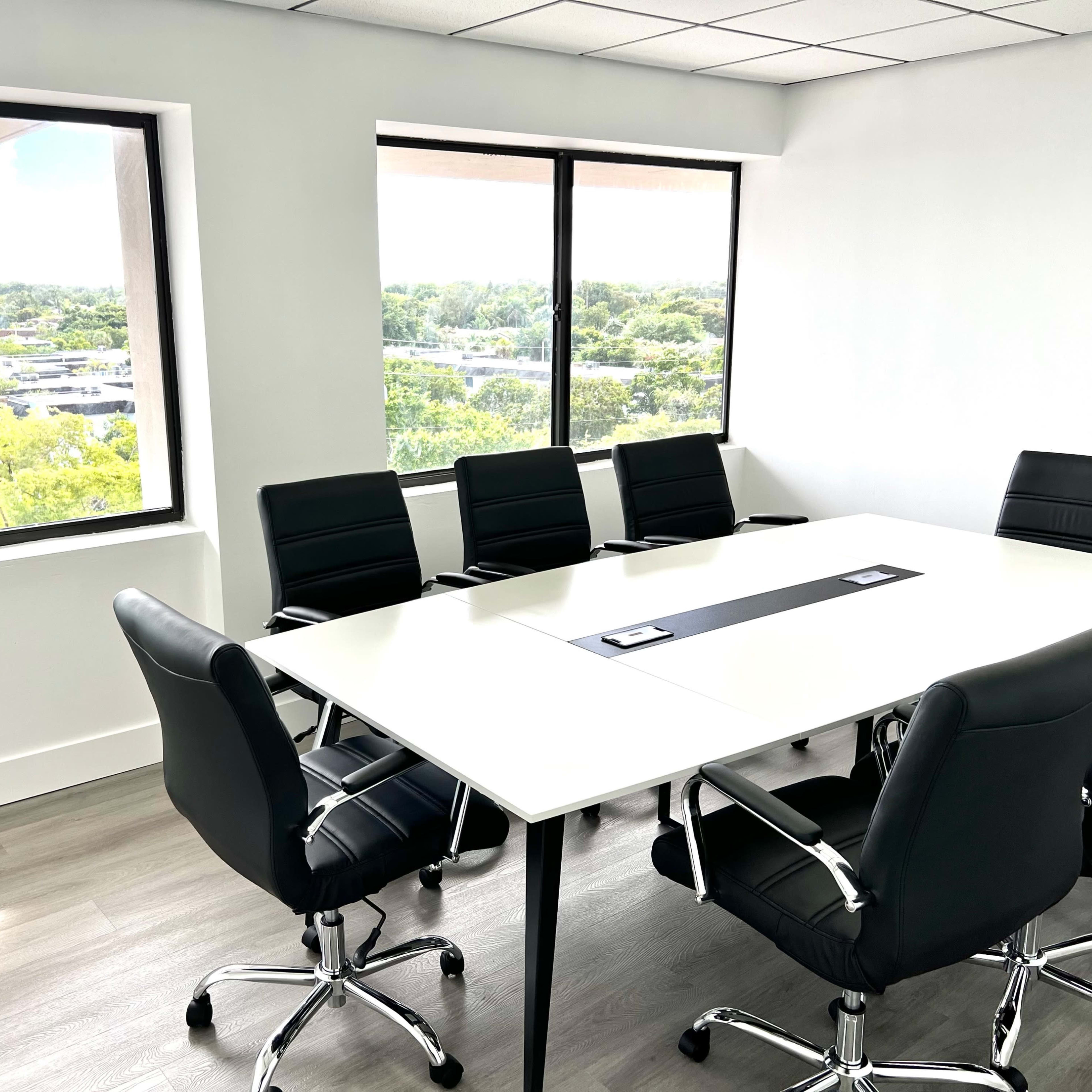 A modern conference room features a large white table surrounded by black leather chairs, with windows offering a view of greenery and a parking lot outside.