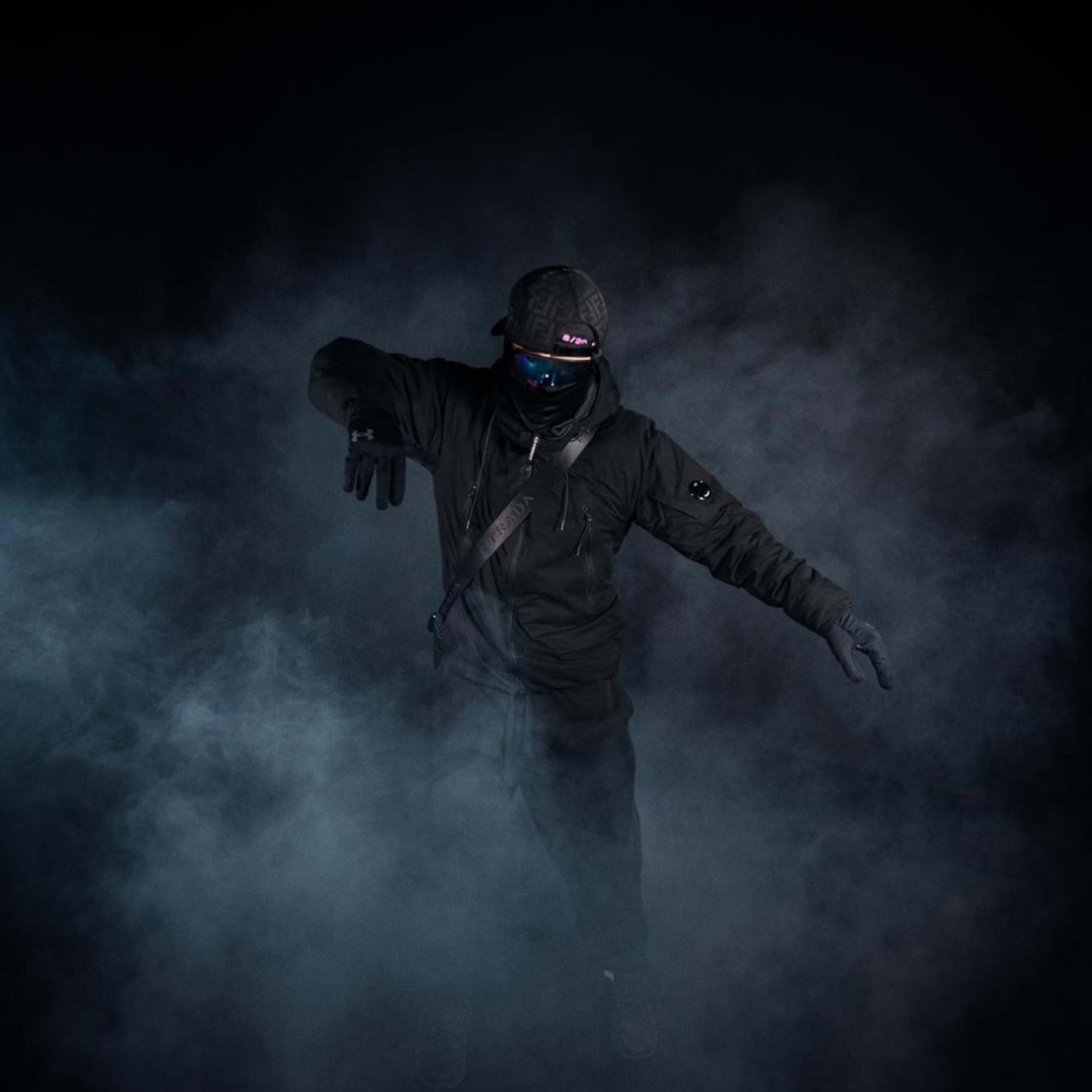 A figure dressed in dark clothing and gloves is posed amidst a dense, smoky background.