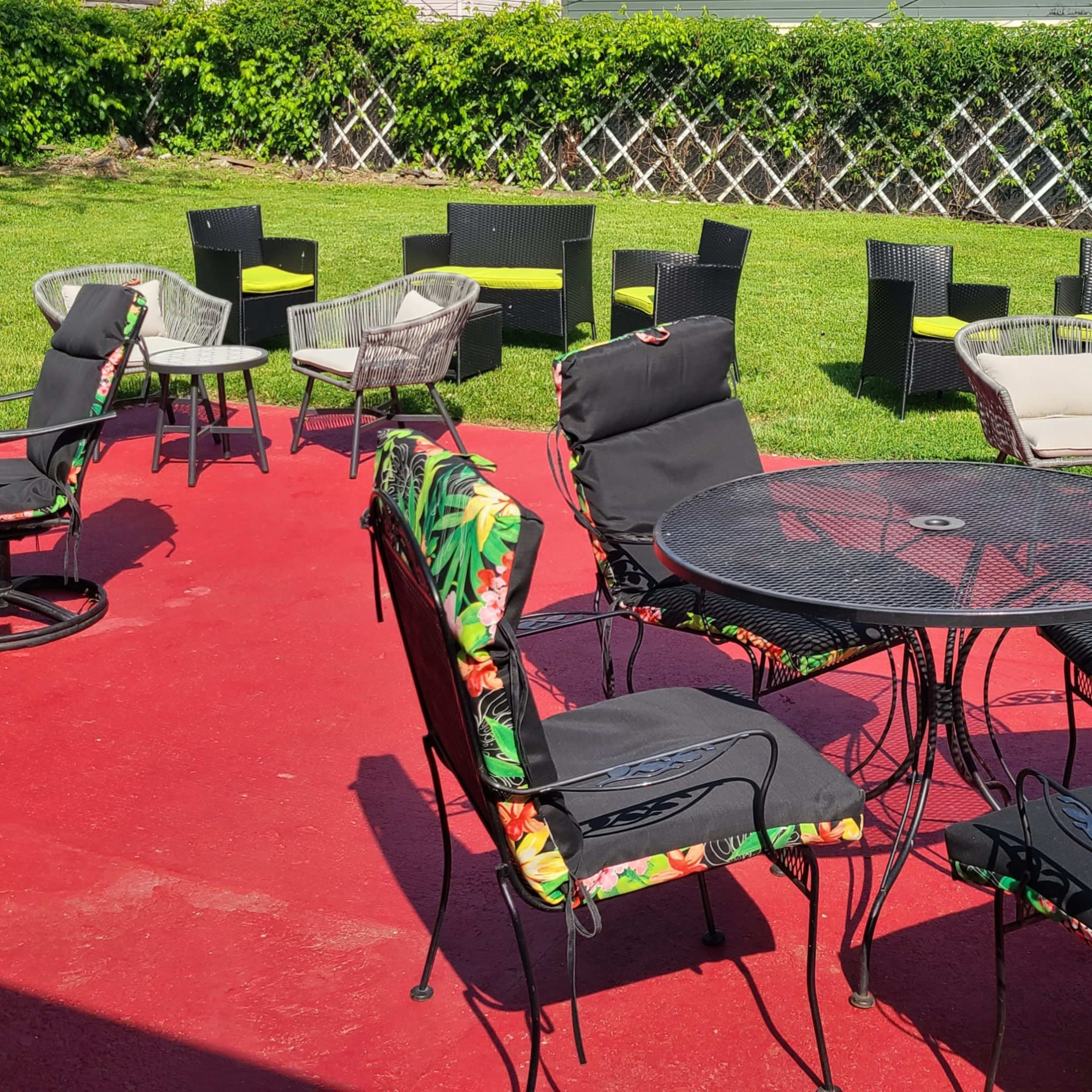 A patio area with various styles of outdoor chairs and tables arranged on a red surface, surrounded by green grass and a vine-covered fence.