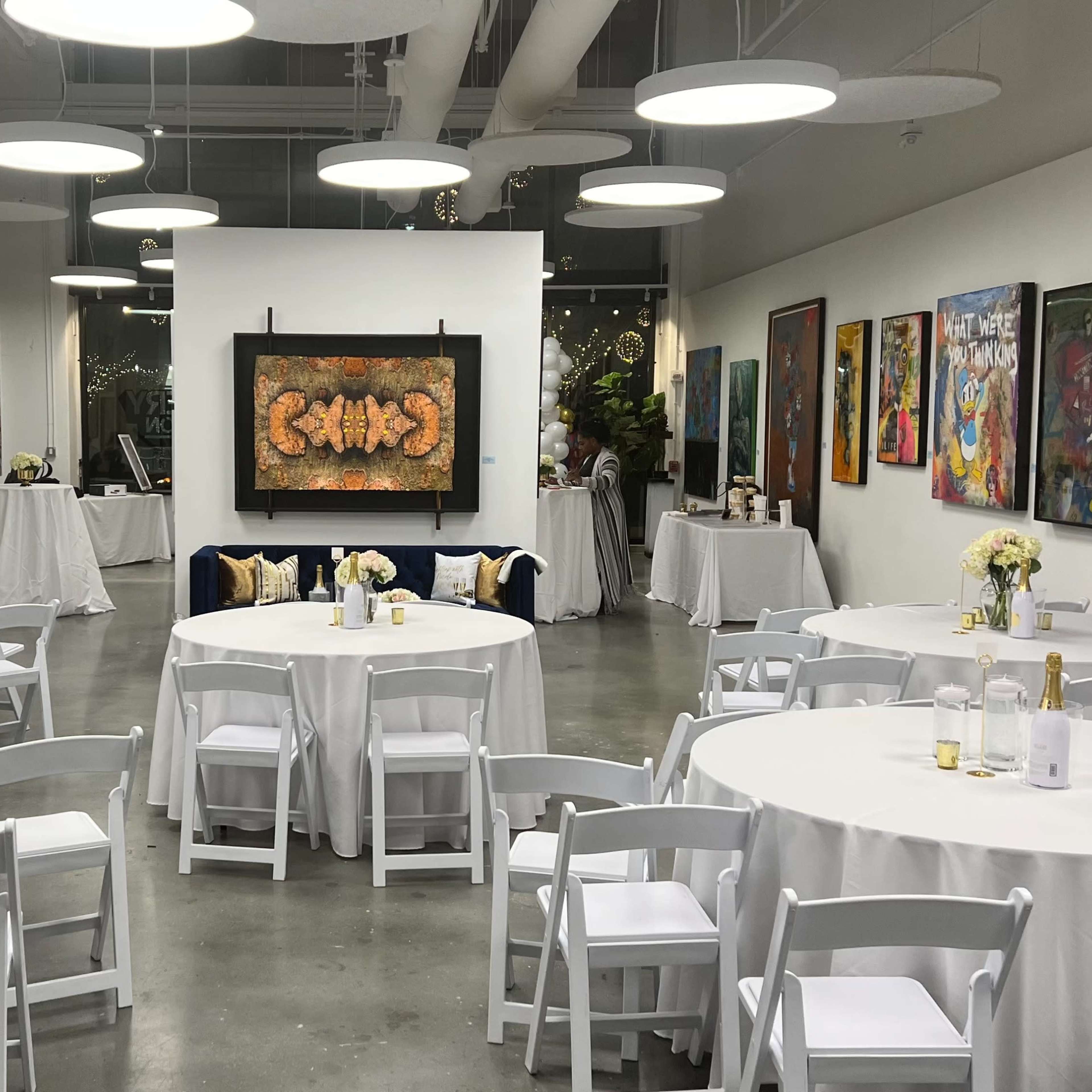 A gallery-style room set up for an event, featuring several round tables covered with white tablecloths, surrounded by artwork hanging on the walls.