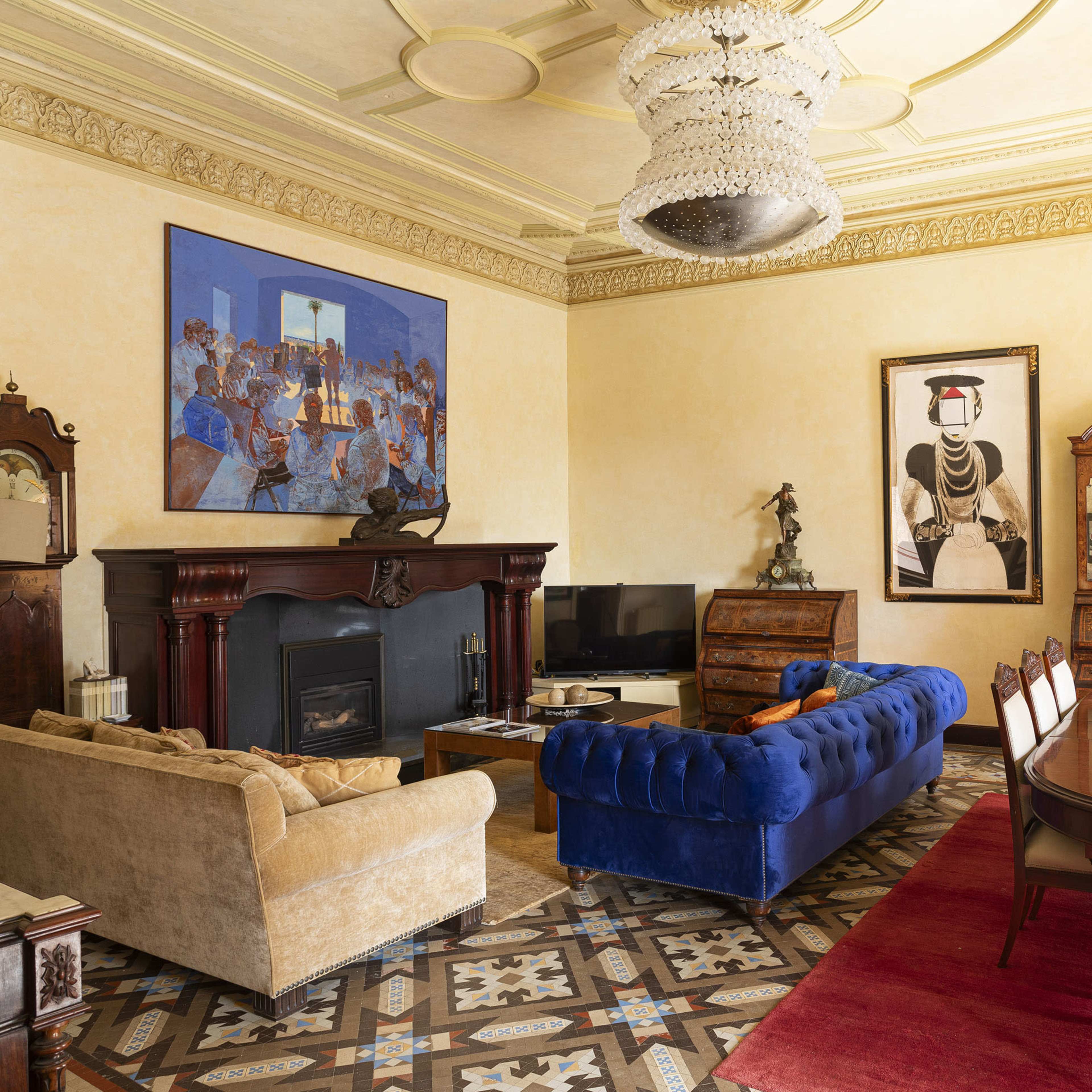 The room features a blue velvet sofa, a beige couch, a wooden dining table, and various artworks on the walls, with a chandelier above and patterned flooring.