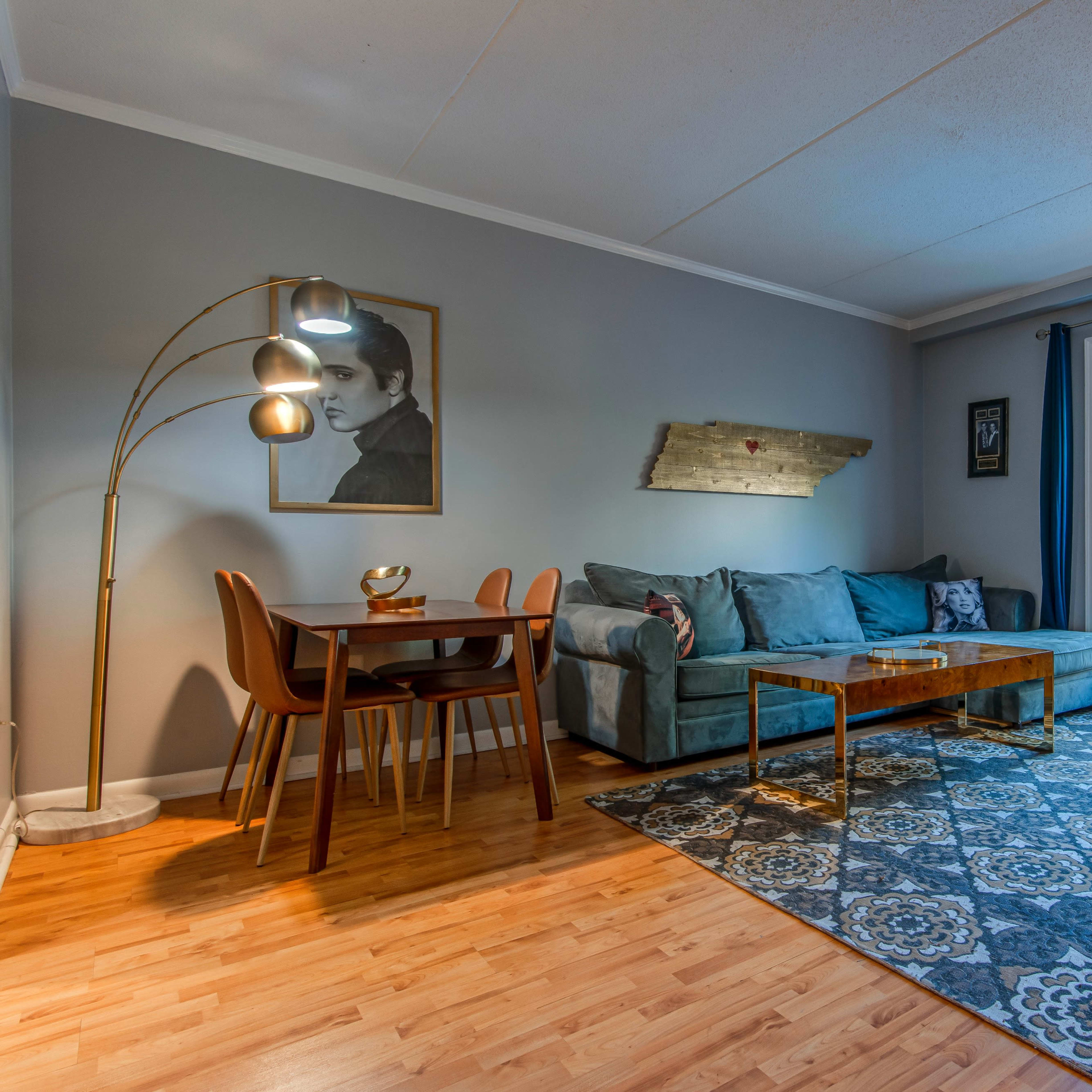 A living room with a teal sofa, a wooden coffee table, a dining area with chairs, and a large window with natural light.